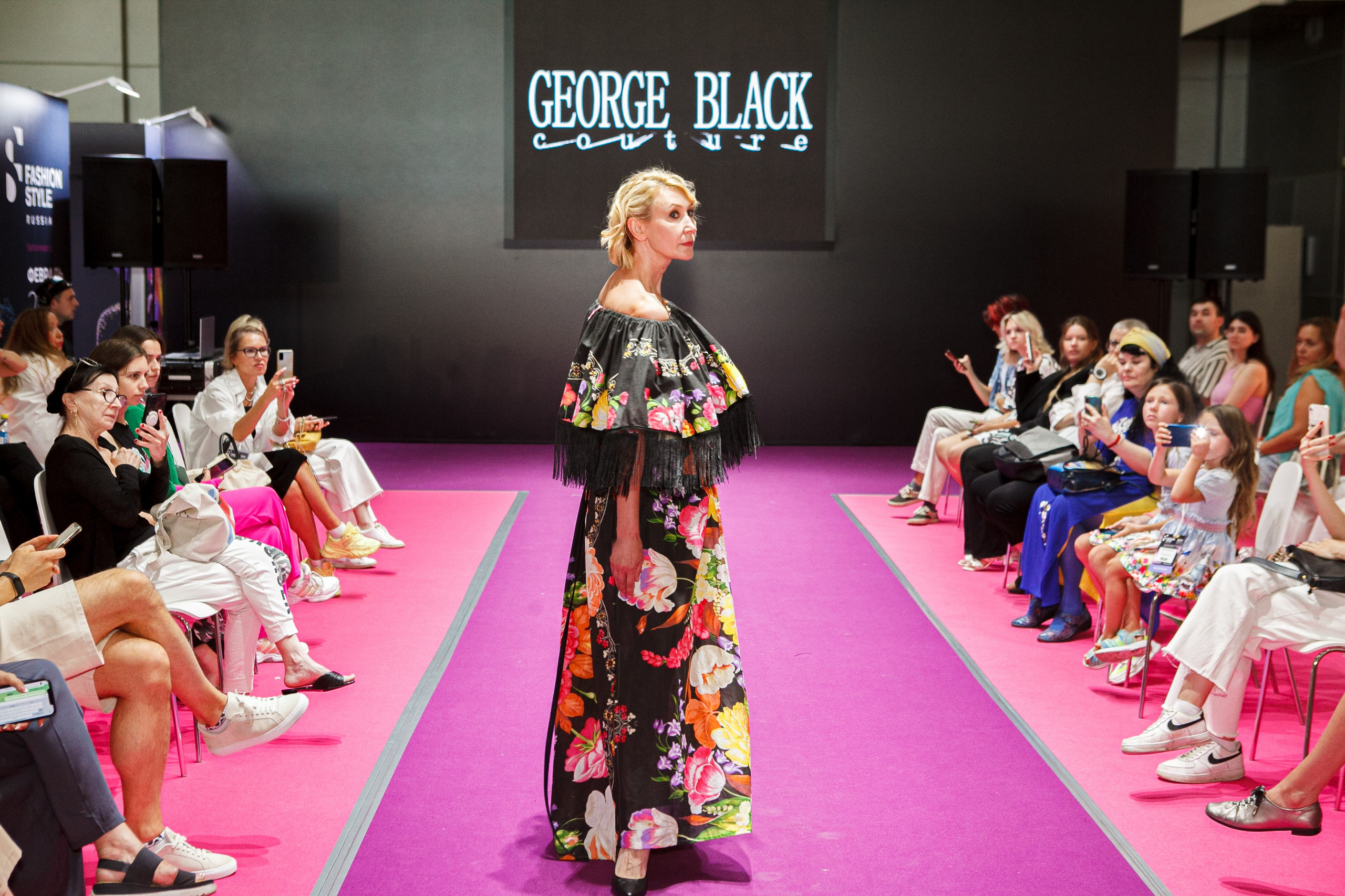  "Модели в ярких нарядах проходят по подиуму во время показа мод, освещенные яркими софитами и окруженные восторженными зрителями."