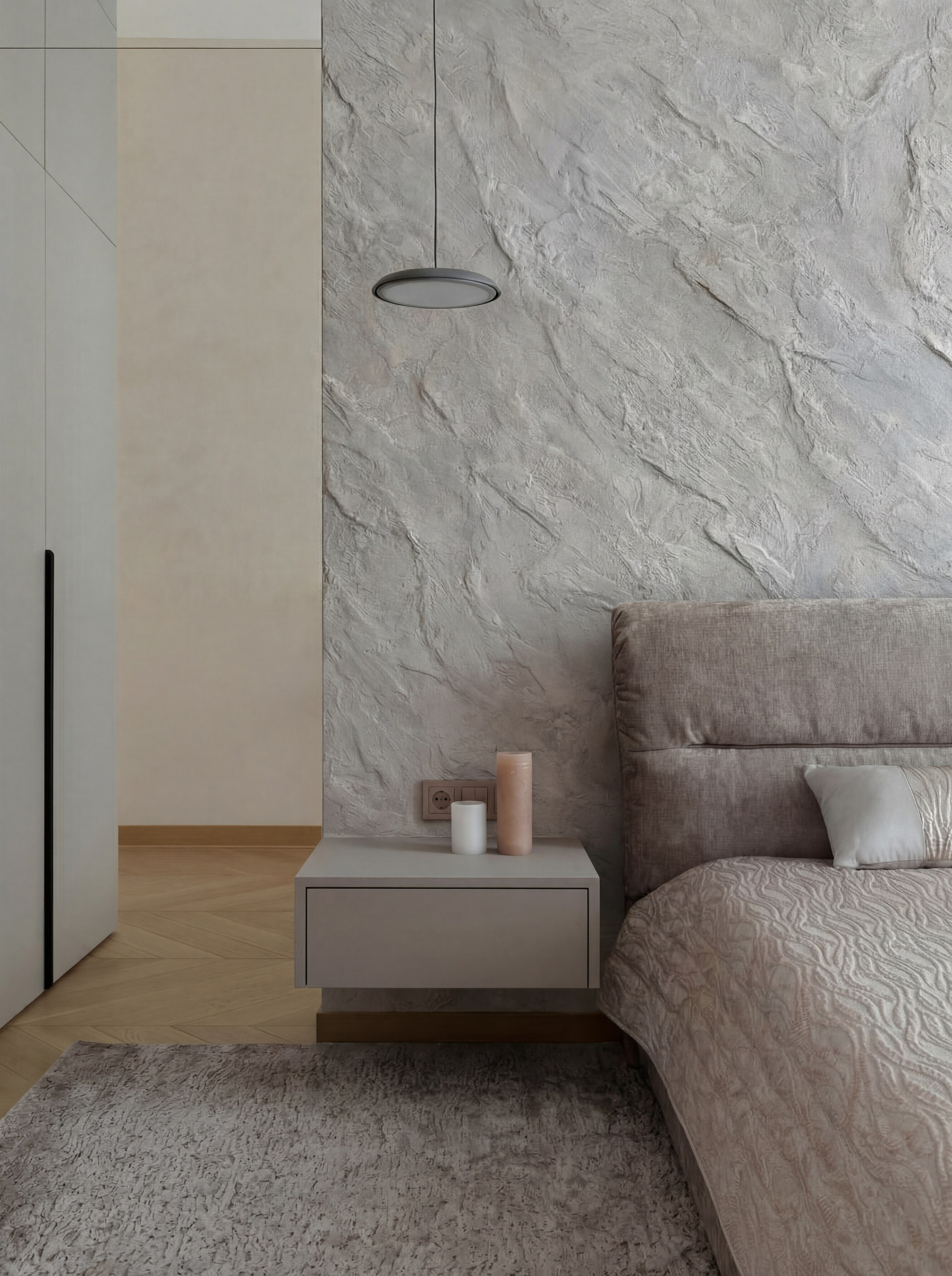 Детальный вид спальни от Валерии Котович: акцентная стена с фактурой камня, подвесная минималистичная тумба, современный светильник и кровать с мягким изголовьем в нейтральных тонах.