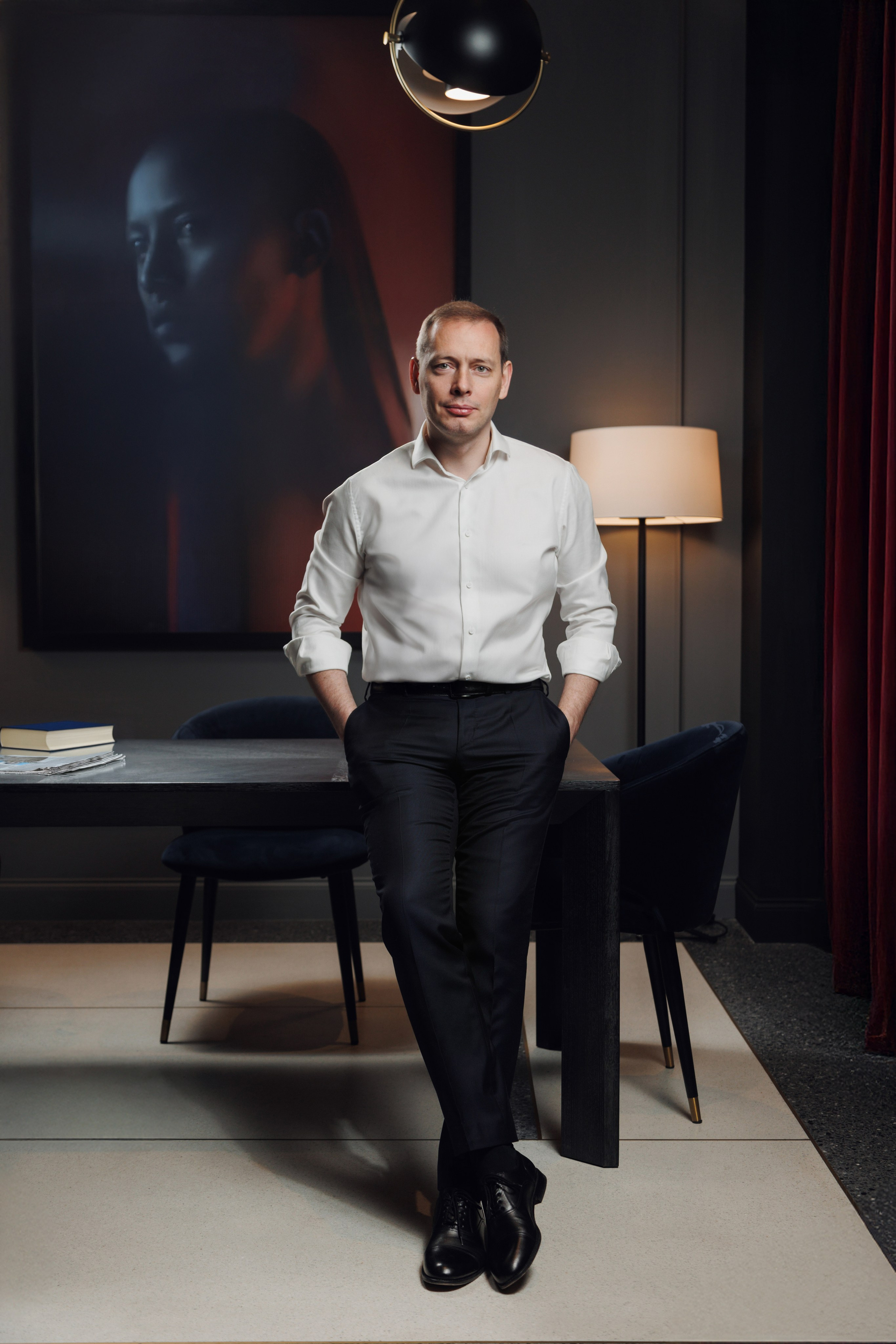 Мужчина в белой рубашке во весь рост на фоне роскошного интерьера отеля.