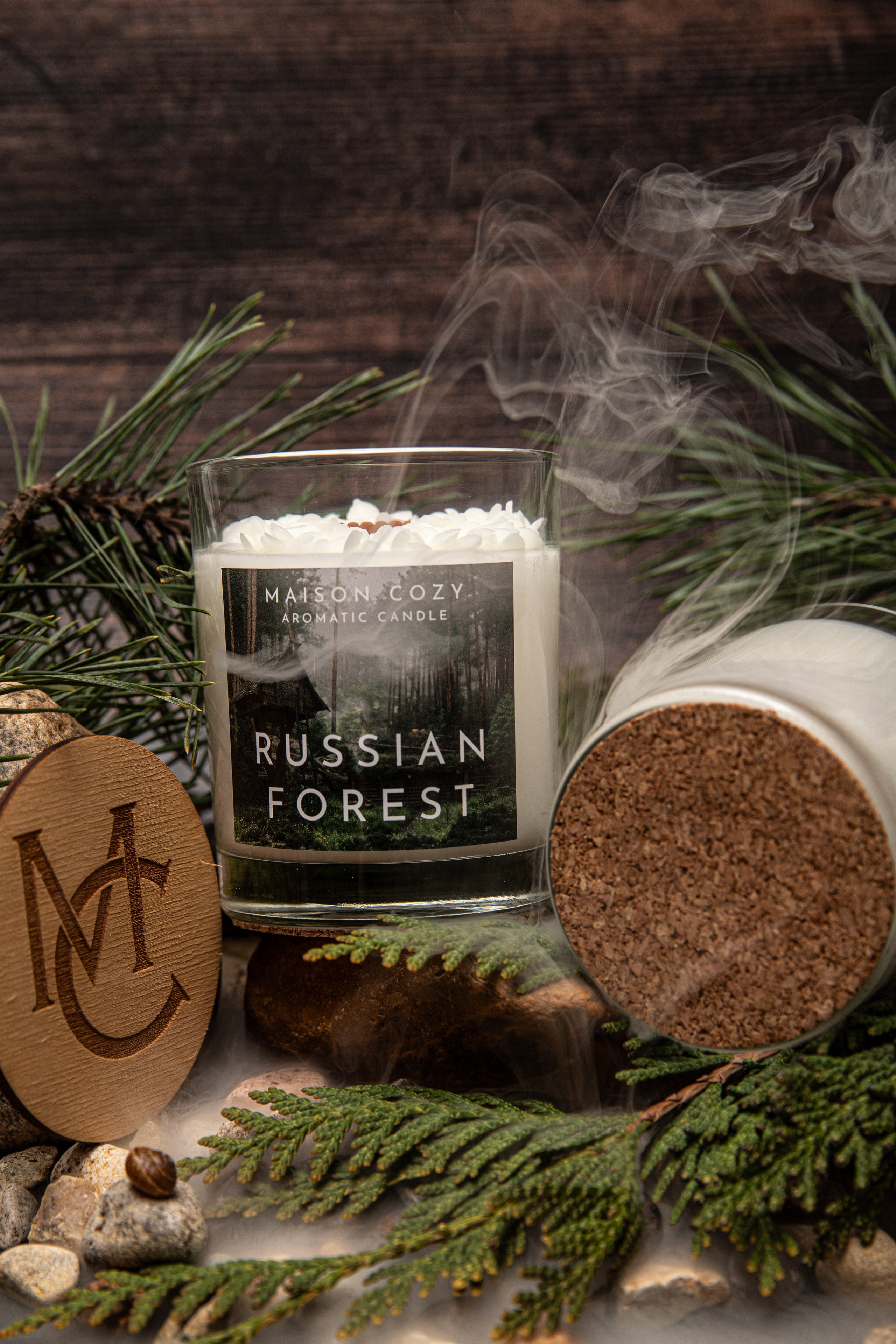 Ароматическая свеча “Русский лес” в прозрачном стеклянном стакане. Свеча окружена натуральной композицией: серыми камнями, ветками сосны и туи с зелёными хвоинками, а также мягким ковром из мха. Фон передаёт атмосферу хвойного леса — свежий, древесный аромат дополнен уютом природного убранства.