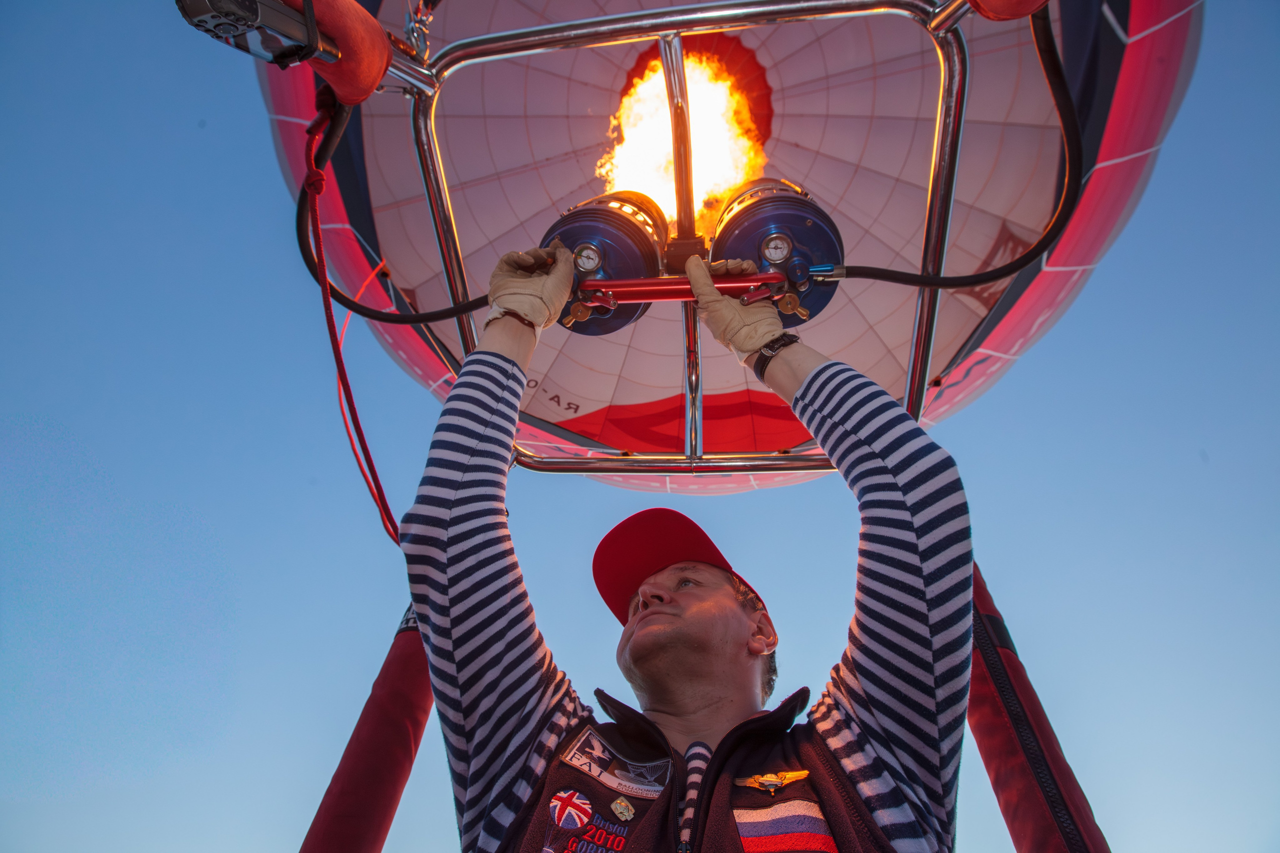 Hot air balloon pilot adjusting the burner system, flame visible above, striped shirt and gloves, clear sky, action moment