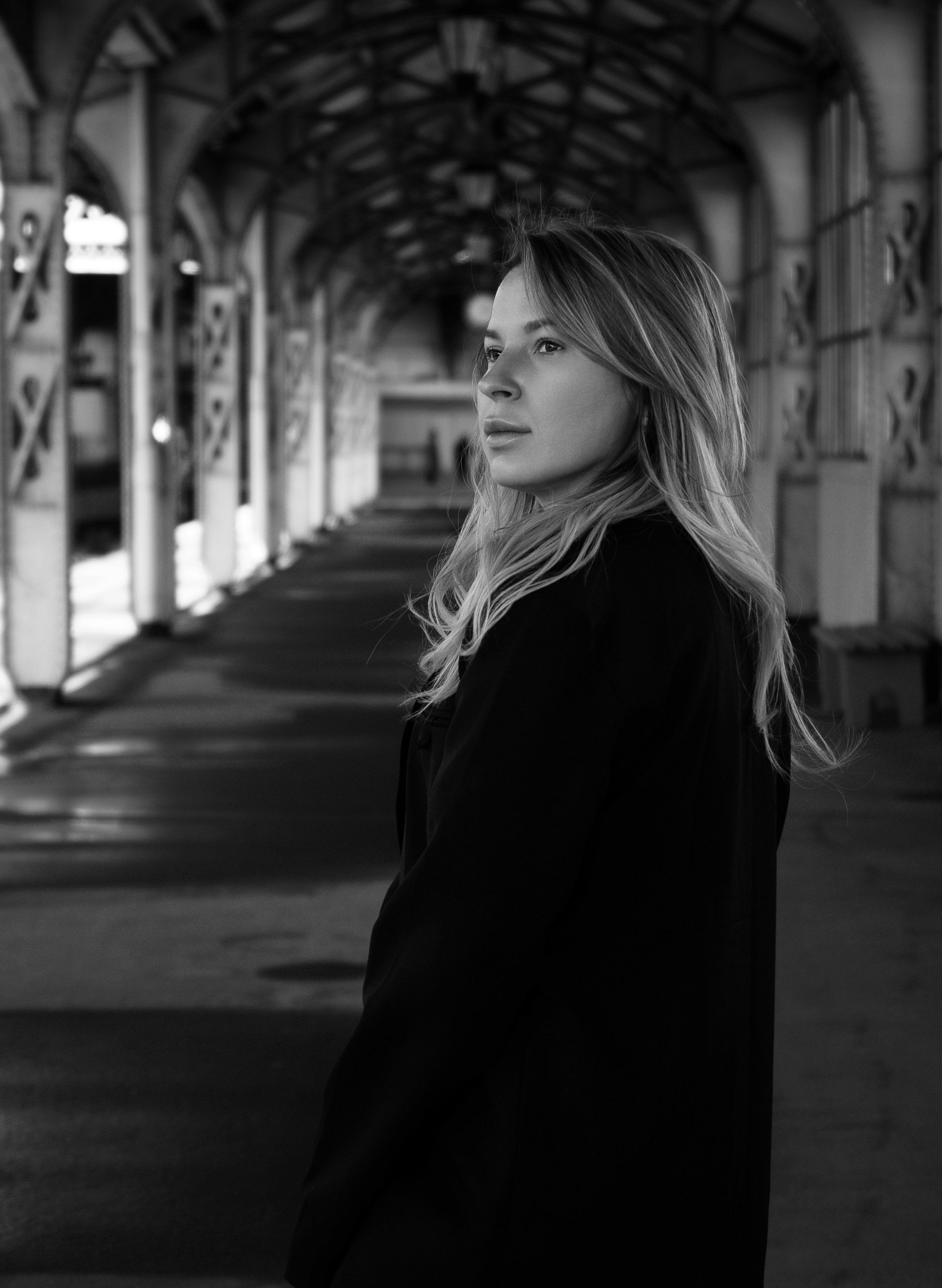 Витебский вокзал. Молодая женщина с длинными светлыми волосами в чёрном пальто стоит в профиль в старинном крытом переходе с арками, чёрно-белое фото.