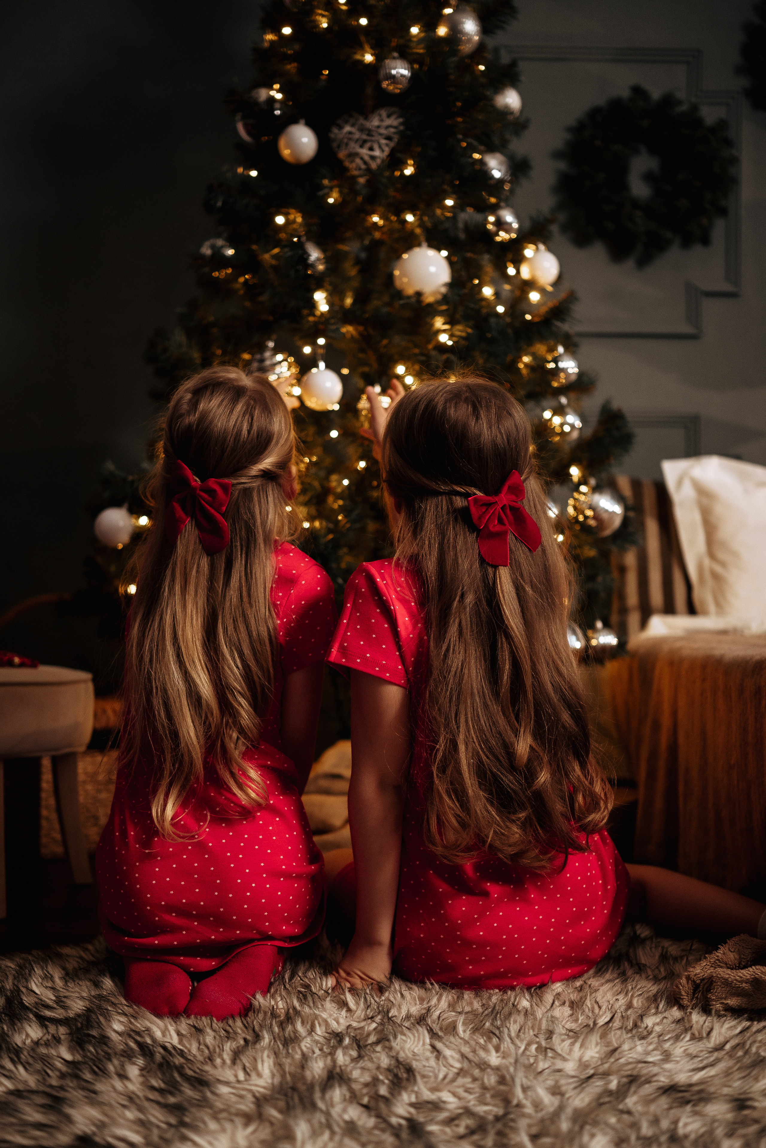 две девочки в красных платьях и с красными бантами в волосах сидят у большой ёлки