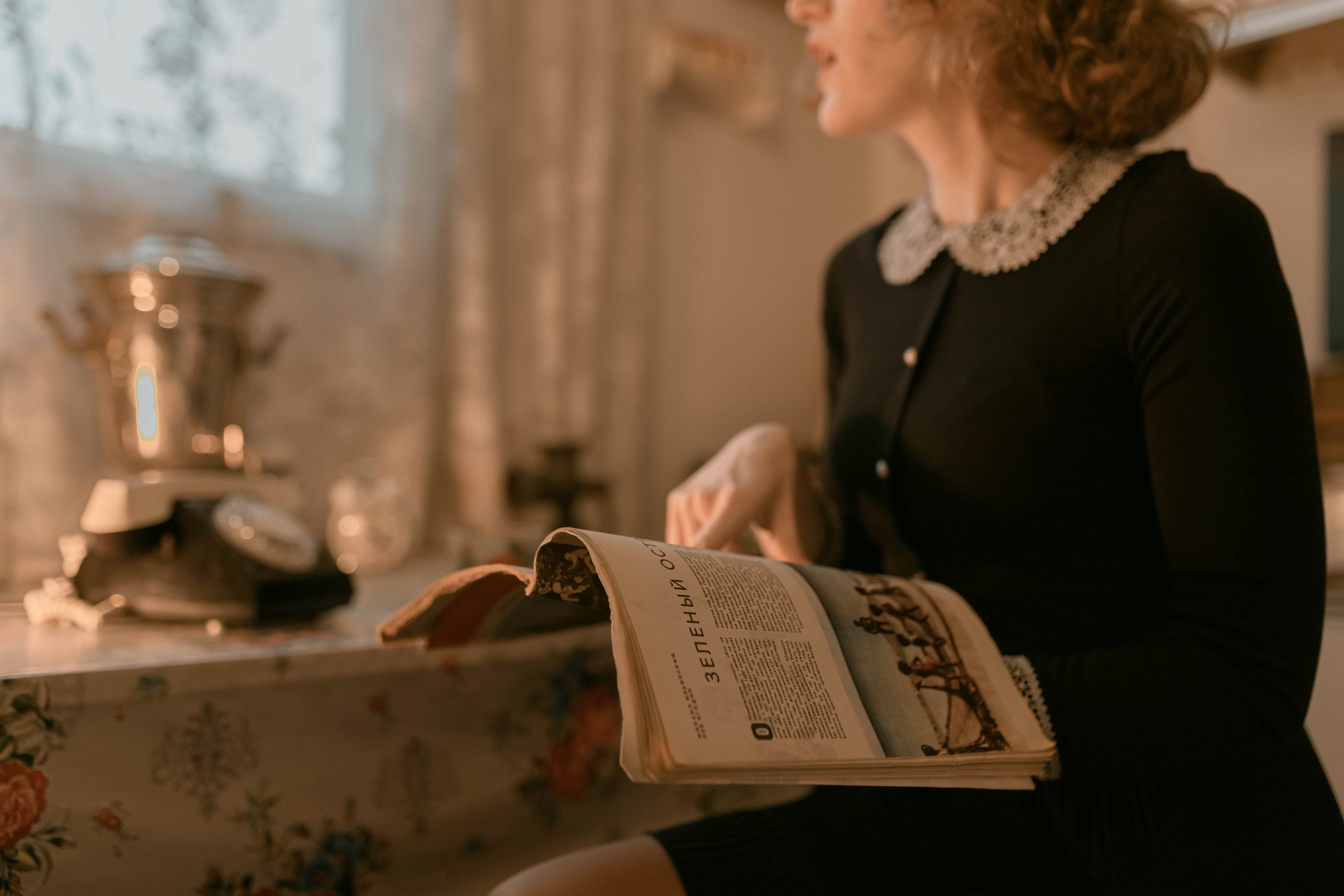 девушка, одетая в классический наряд, сидит за столом, покрытым цветочным узором, и читает книгу. Зал залит теплым естественным светом, что создает уютную атмосферу.