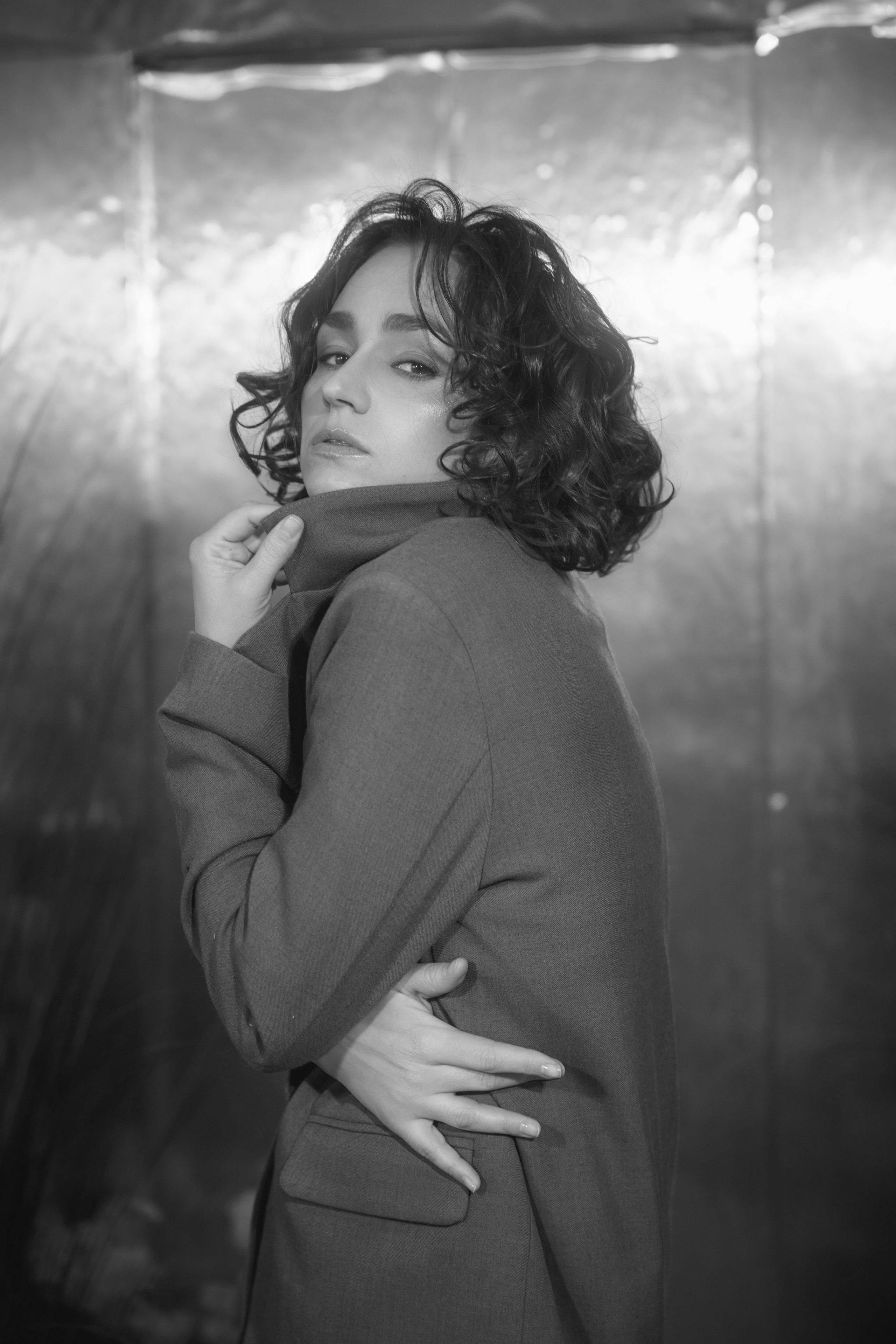 Портретное фото молодой девушки в студии. Девушка сидит на фоне нейтрального цвета, ее лицо освещено мягким светом. Она смотрит прямо в камеру с легкой улыбкой. На девушке стильная одежда, подчеркивающая современный модный стиль. Фотография выполнена в высоком разрешении, с акцентом на деталях