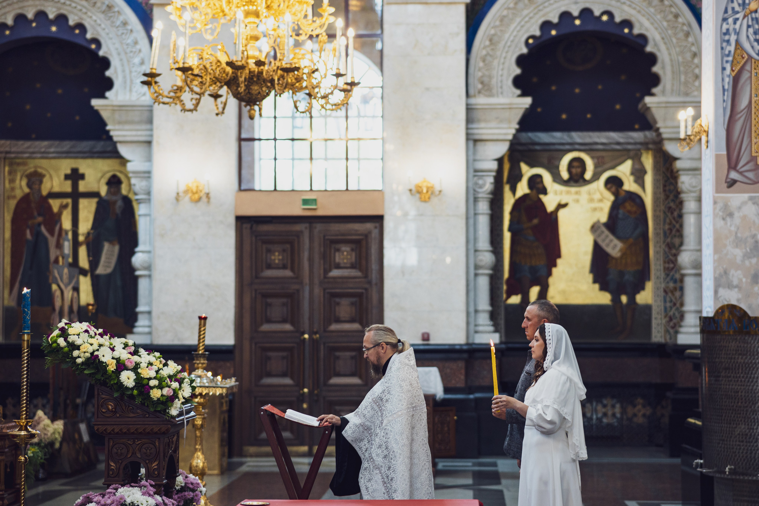 Священник читает молитву у аналоя, пара со свечами, белый иконостас и цветы — Екатеринбург, Илья Антропов