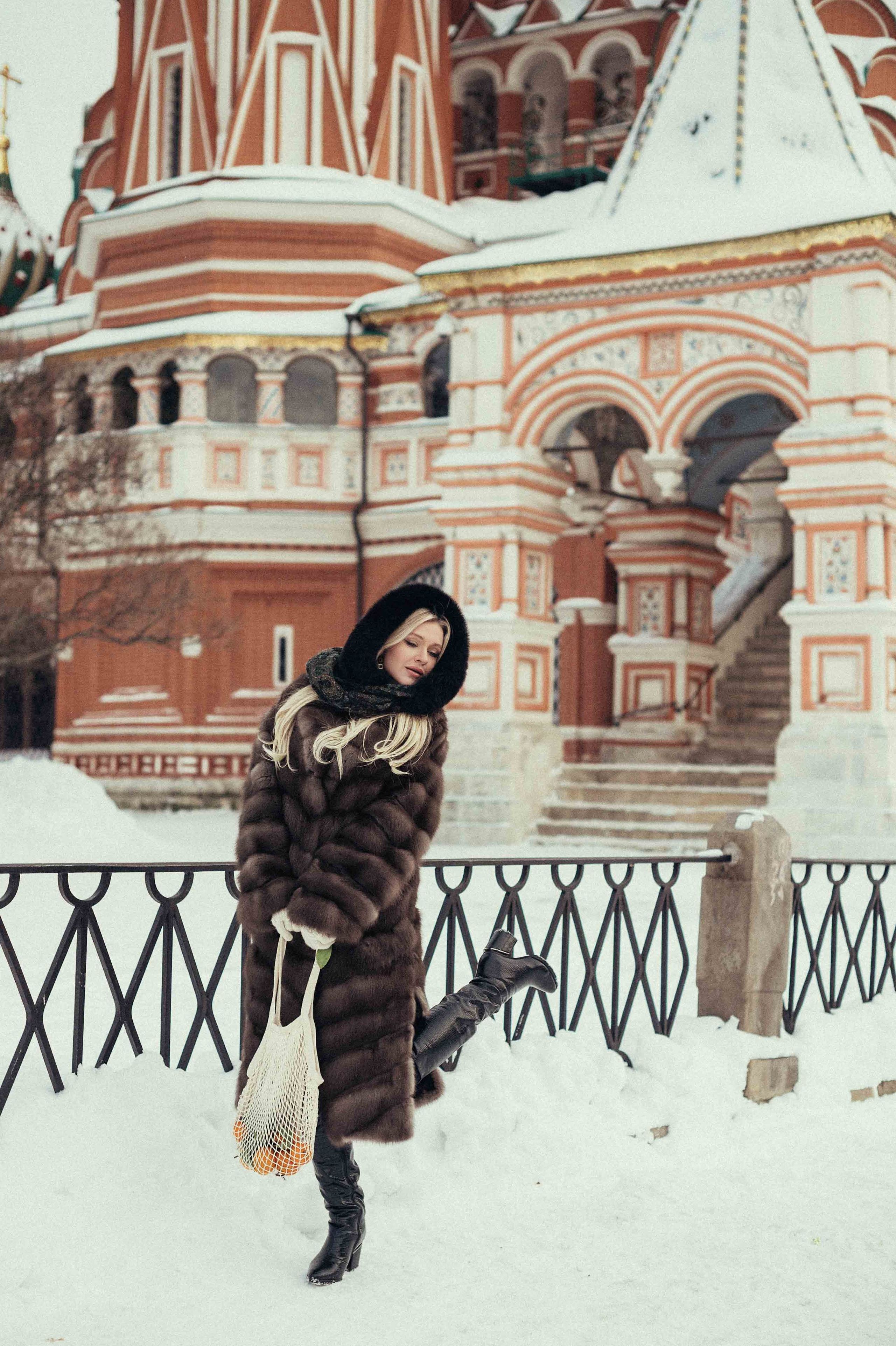 Фотосессия в стиле Russian girl на Красной площади на фоне храма Василия Блаженного, на которой девушка стоит в соболиной шубе, держа авоську мандаринов в руках, и игриво закинула ножку в танце