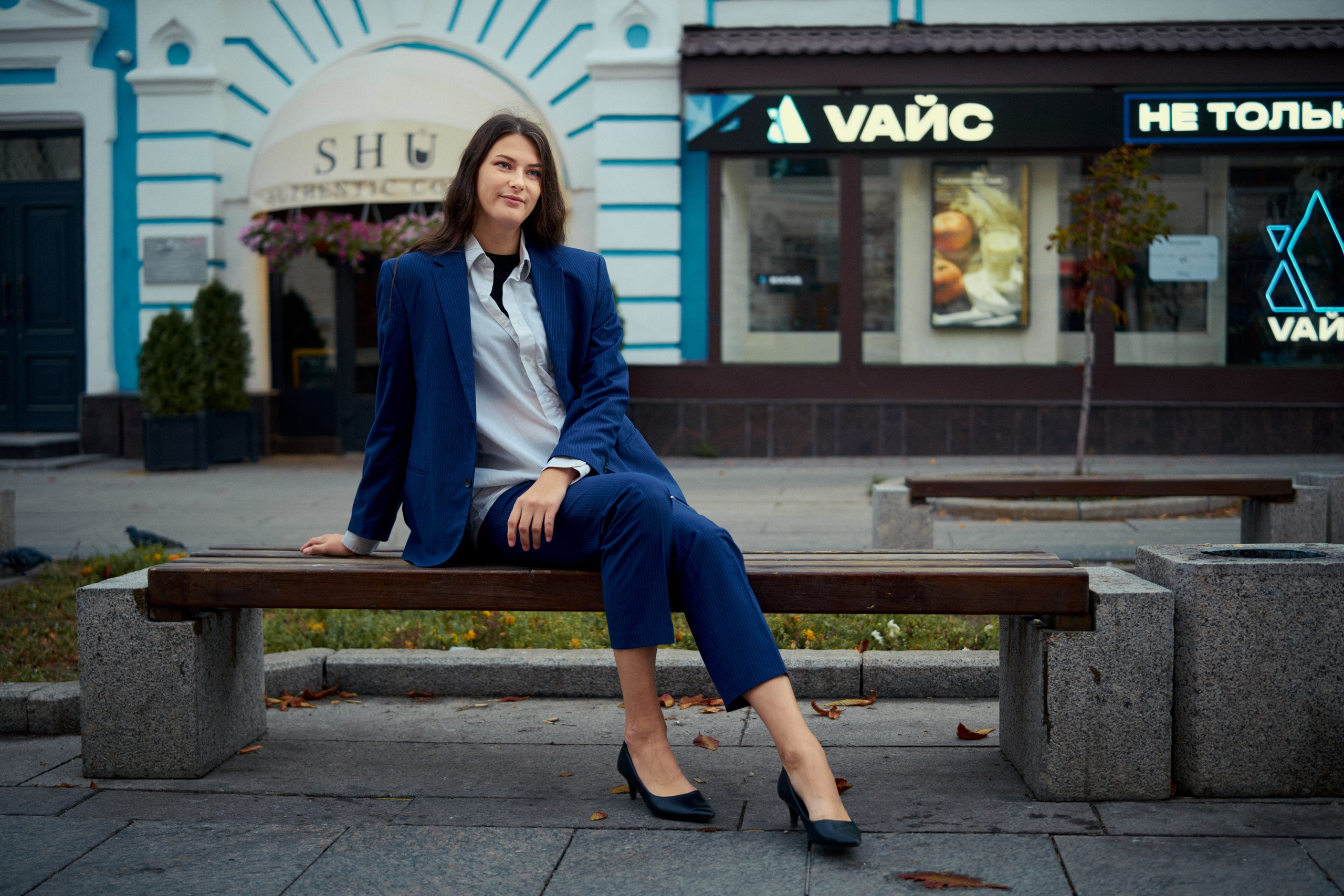 Элегантная девушка в синем брючном костюме и белой рубашке, сидит на фоне городской архитектуры