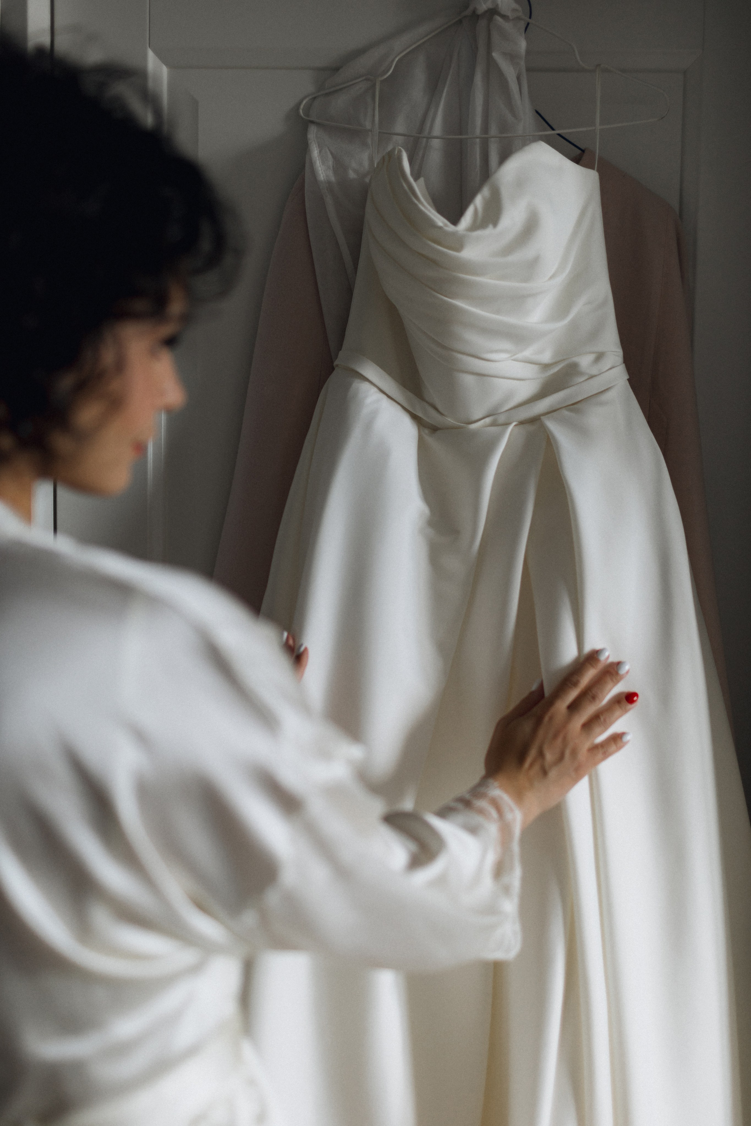 Свадебная фотография в стиле эстетики тишины: невеста, рассматривающая платье на вешалке в интерьере заполненным мягким боковым светом