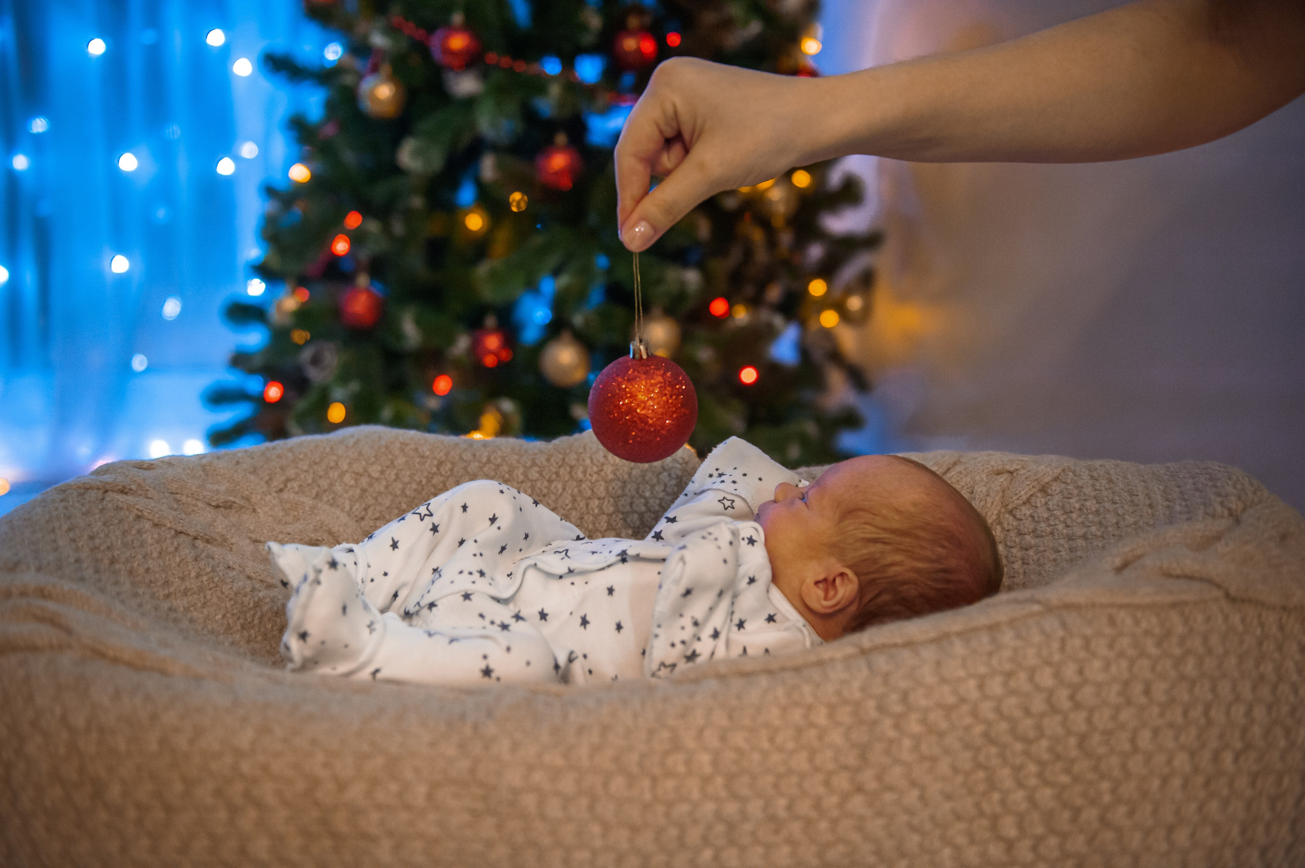 новорожденный ребенок лежит у елки и смотрит на елочную игрушку