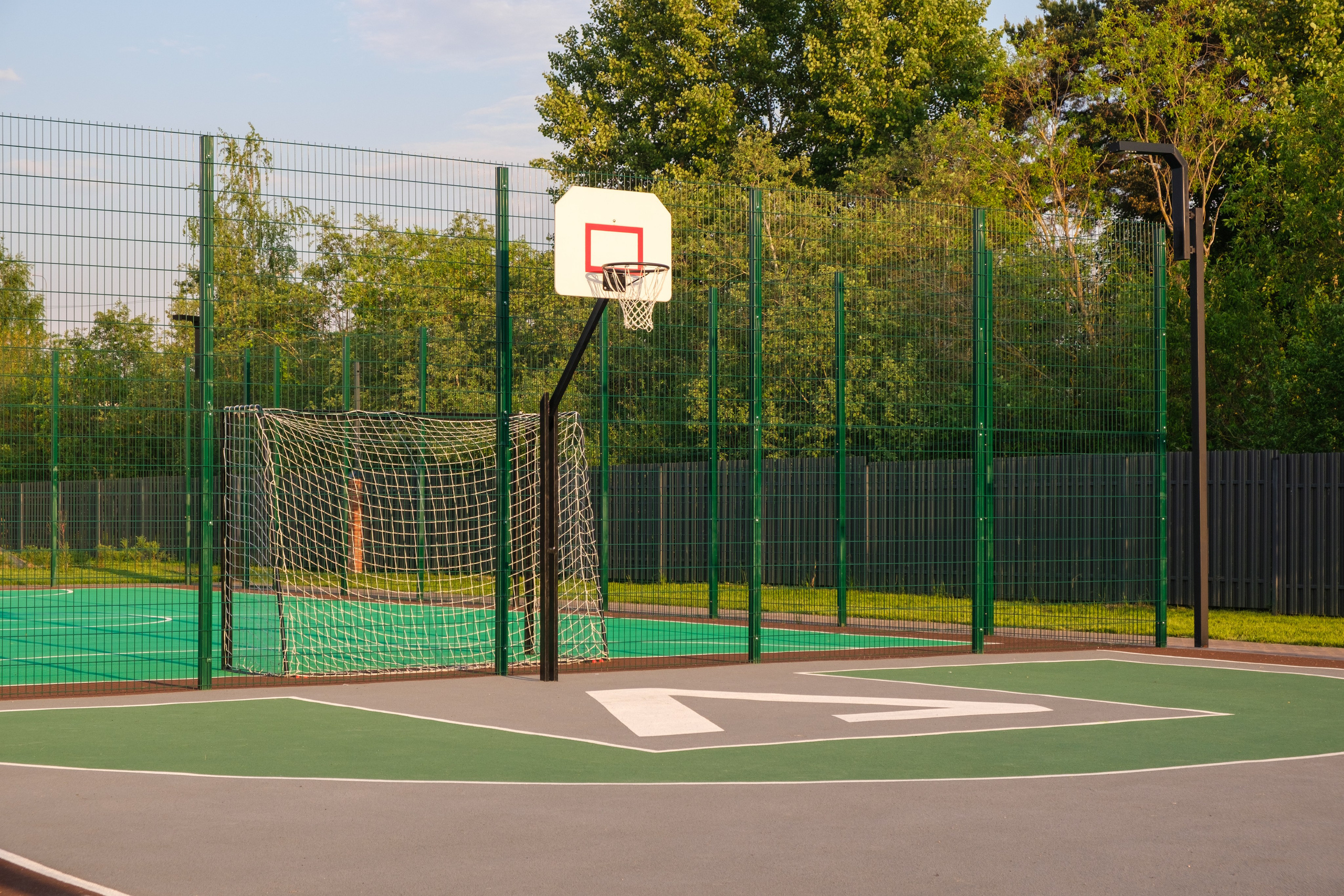 Спортивная площадка с зелёным покрытием для игры в футбол или баскетбол. Видна сетка ворот и баскетбольный щит с кольцом. Площадка окружена высоким забором из металлической сетки.