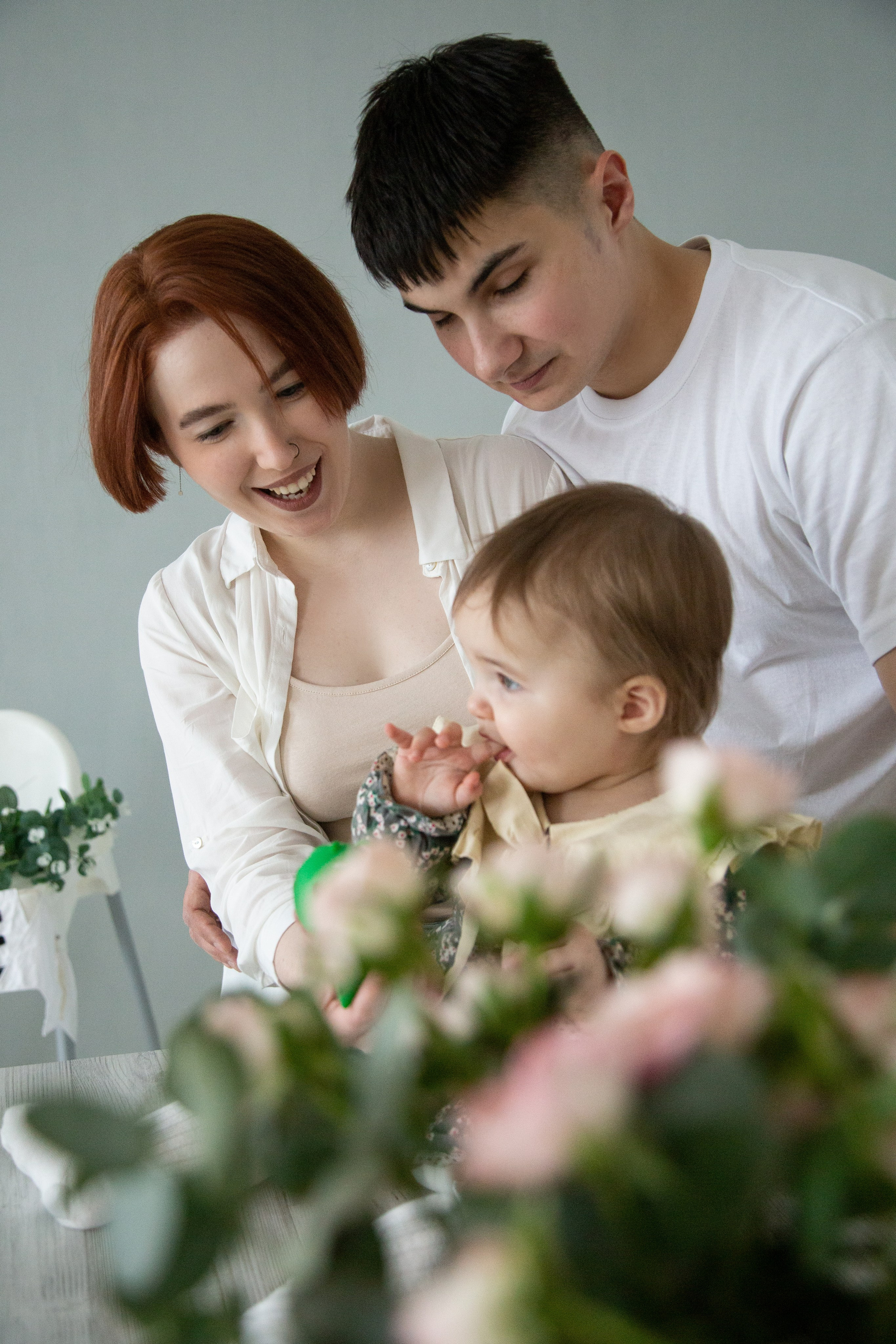 Семейное фото в интерьере, где мать, отец и двое детей обнимаются, сидя на диване, вокруг них растительность и цветы.