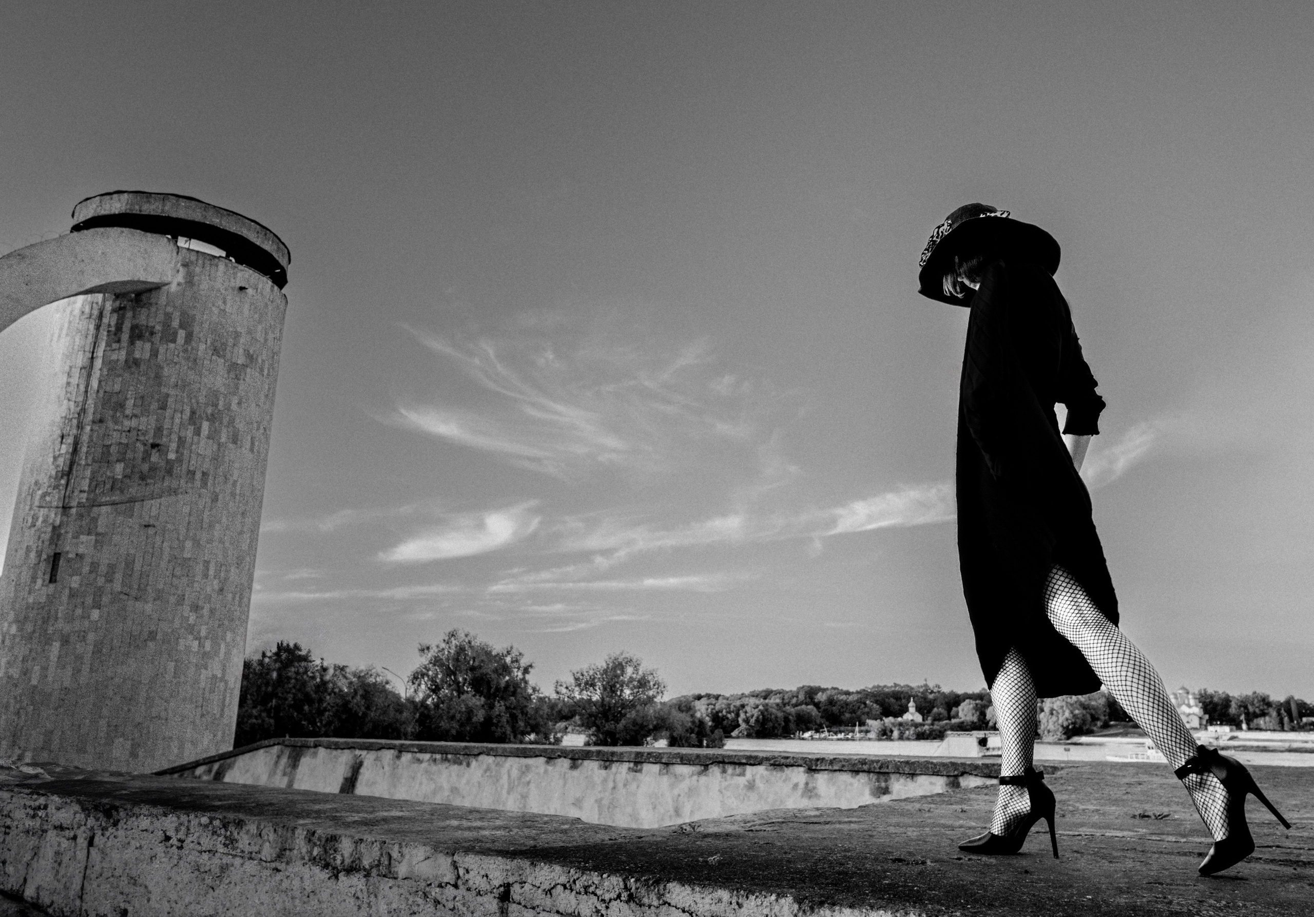 Женщина в чёрном платье и сетчатых колготках идёт по крыше на фоне архитектурной башни и неба, чёрно-белая арт-съёмка