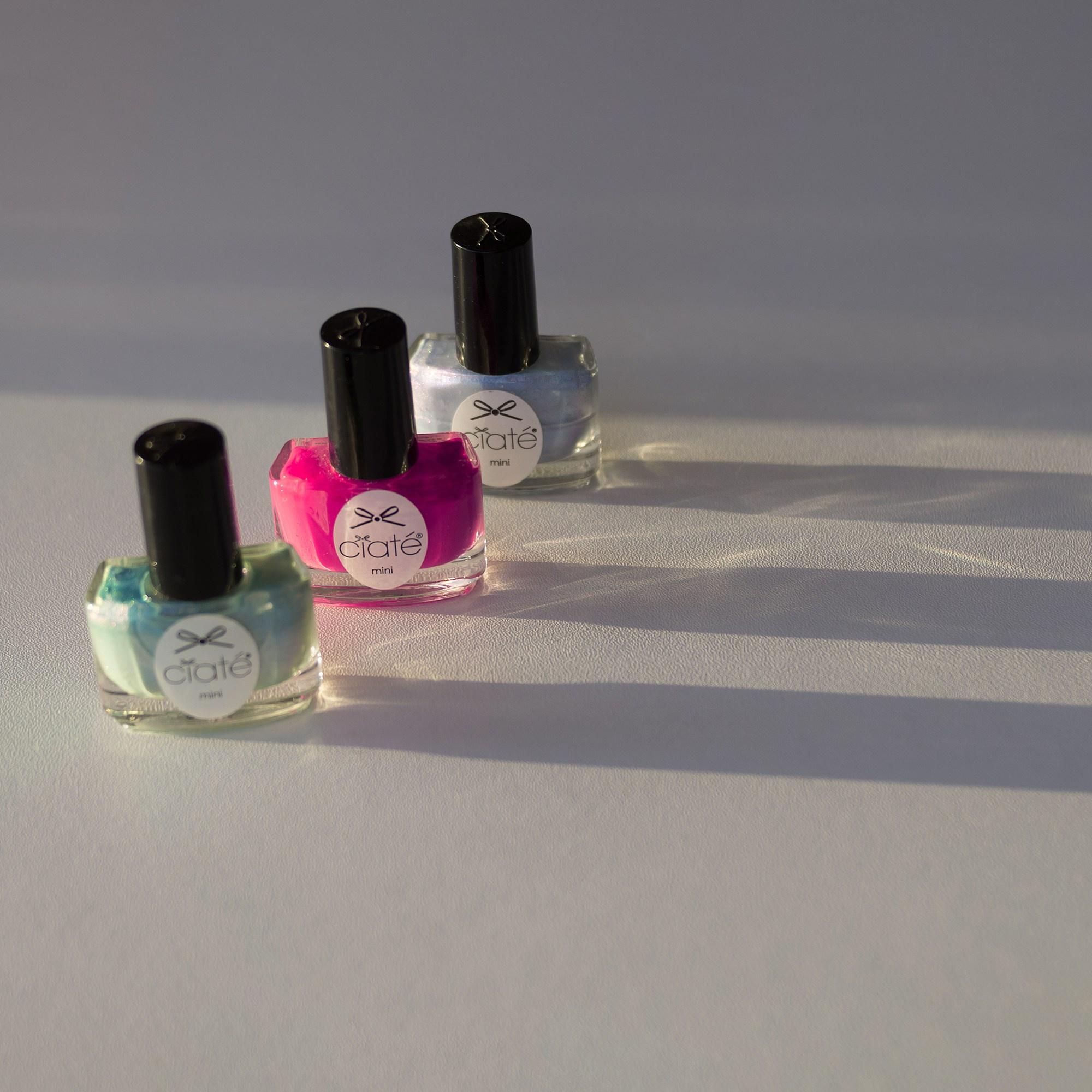 Три флакона с цветным лаком для ногтей на светлой поверхности. Боковое освещение, длинные тени.