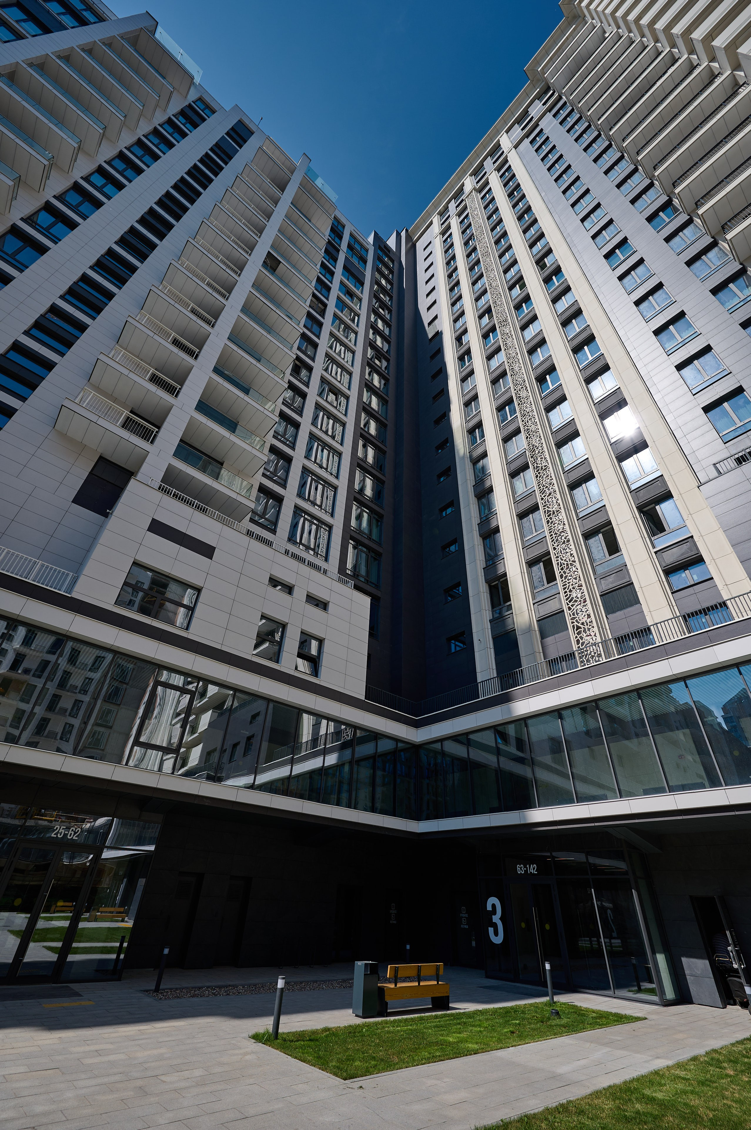 Перспективный вид вверх на фасады высоких жилых зданий, сходящихся под острым углом; внизу — входная группа с зелёной лужайкой и лавочкой