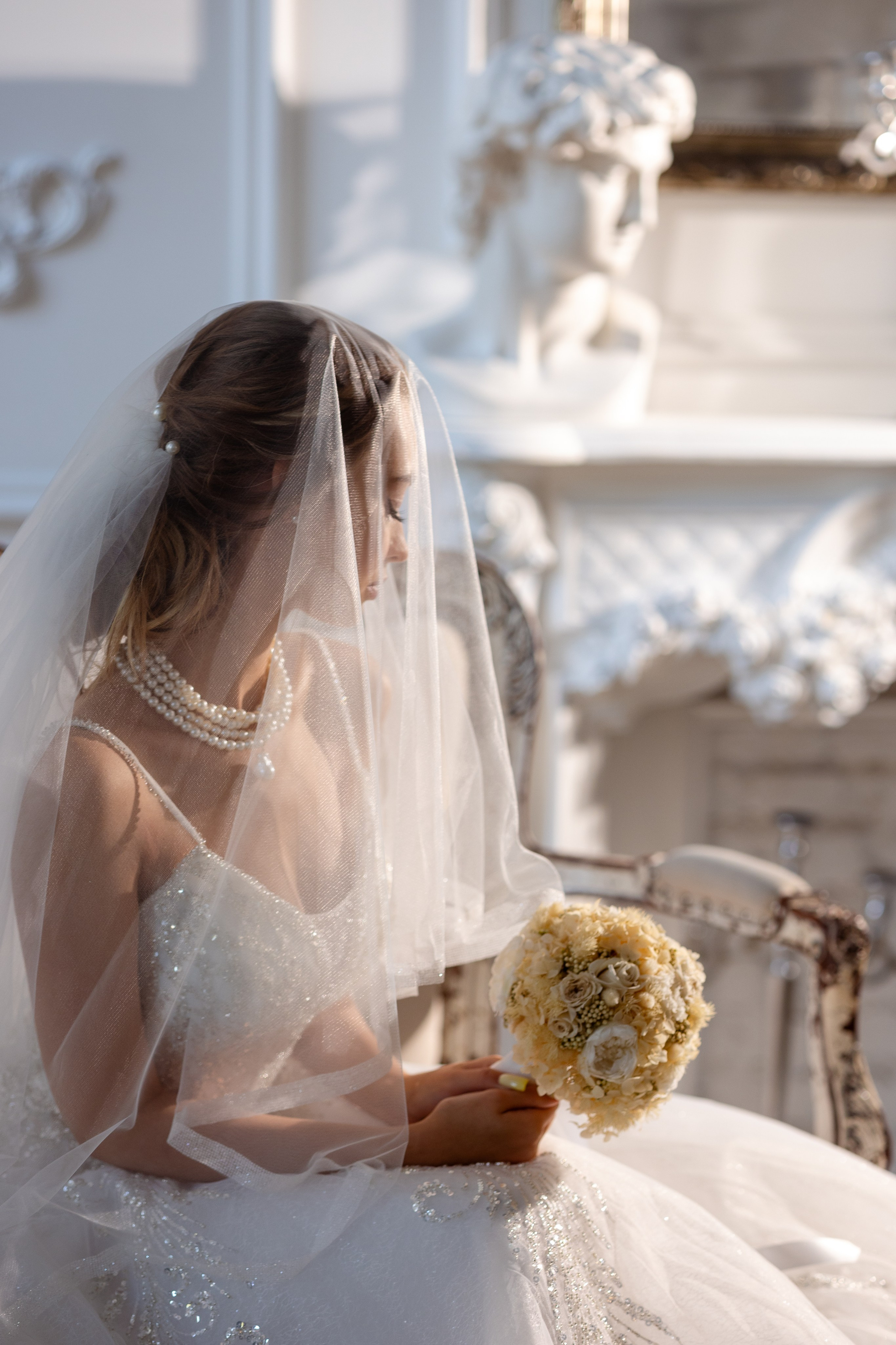 Невеста сидит с букетом в руках, накрытая белоснежной вуалью.