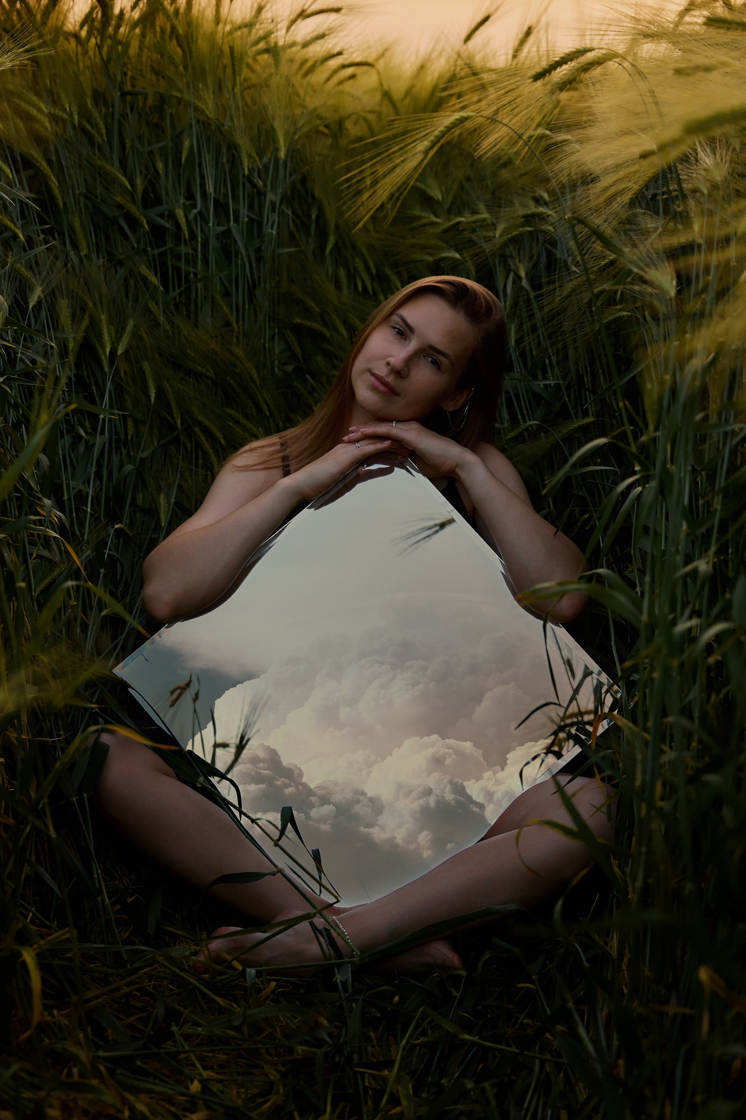 Портрет девушки в зеленом ржаном поле на закате летом. Девушка сидя на траве обнимает зеркало. В зеркале отражается небо с облаками. Индивидуальные фотосессии в Анапе.