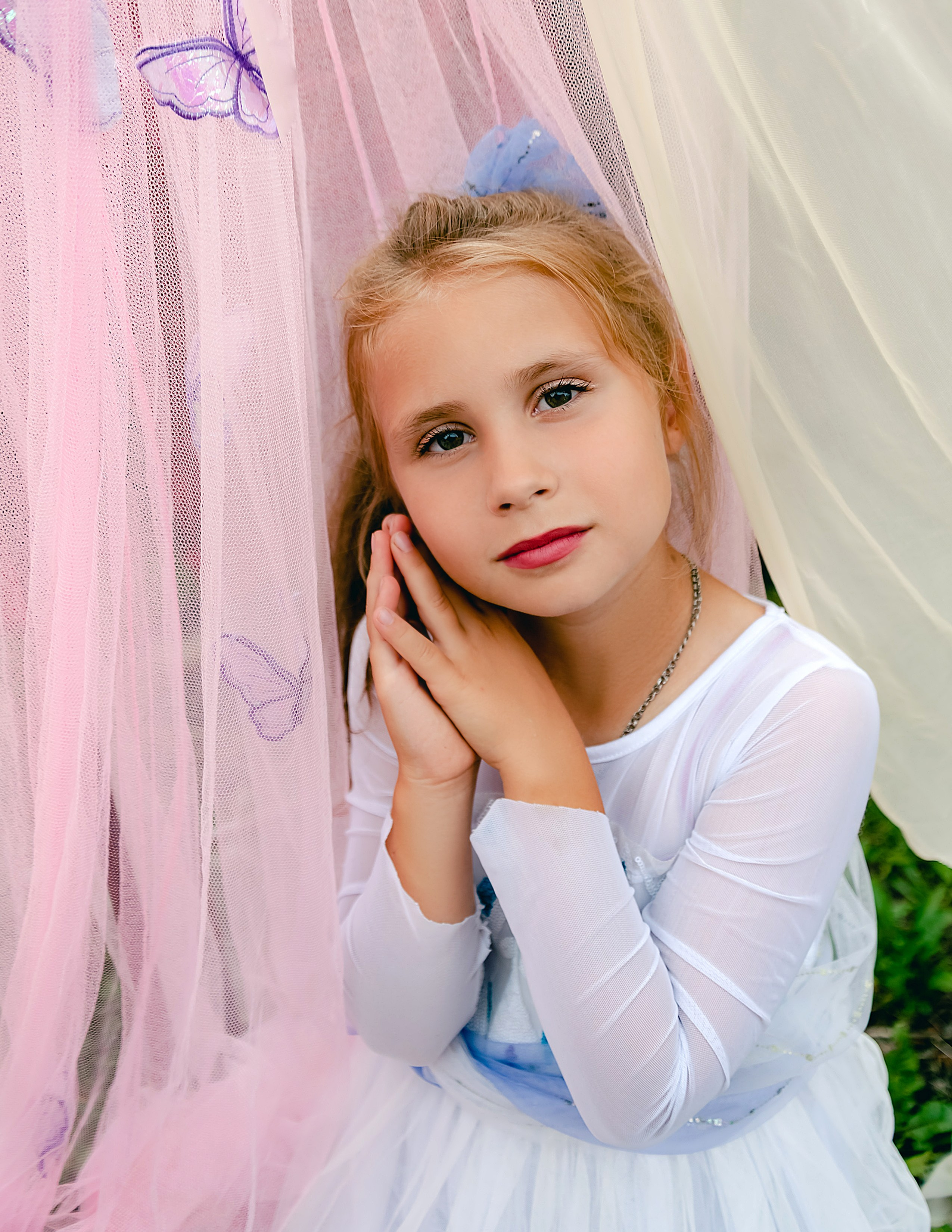 На фото милая девочка сидит и держит руки у лица.