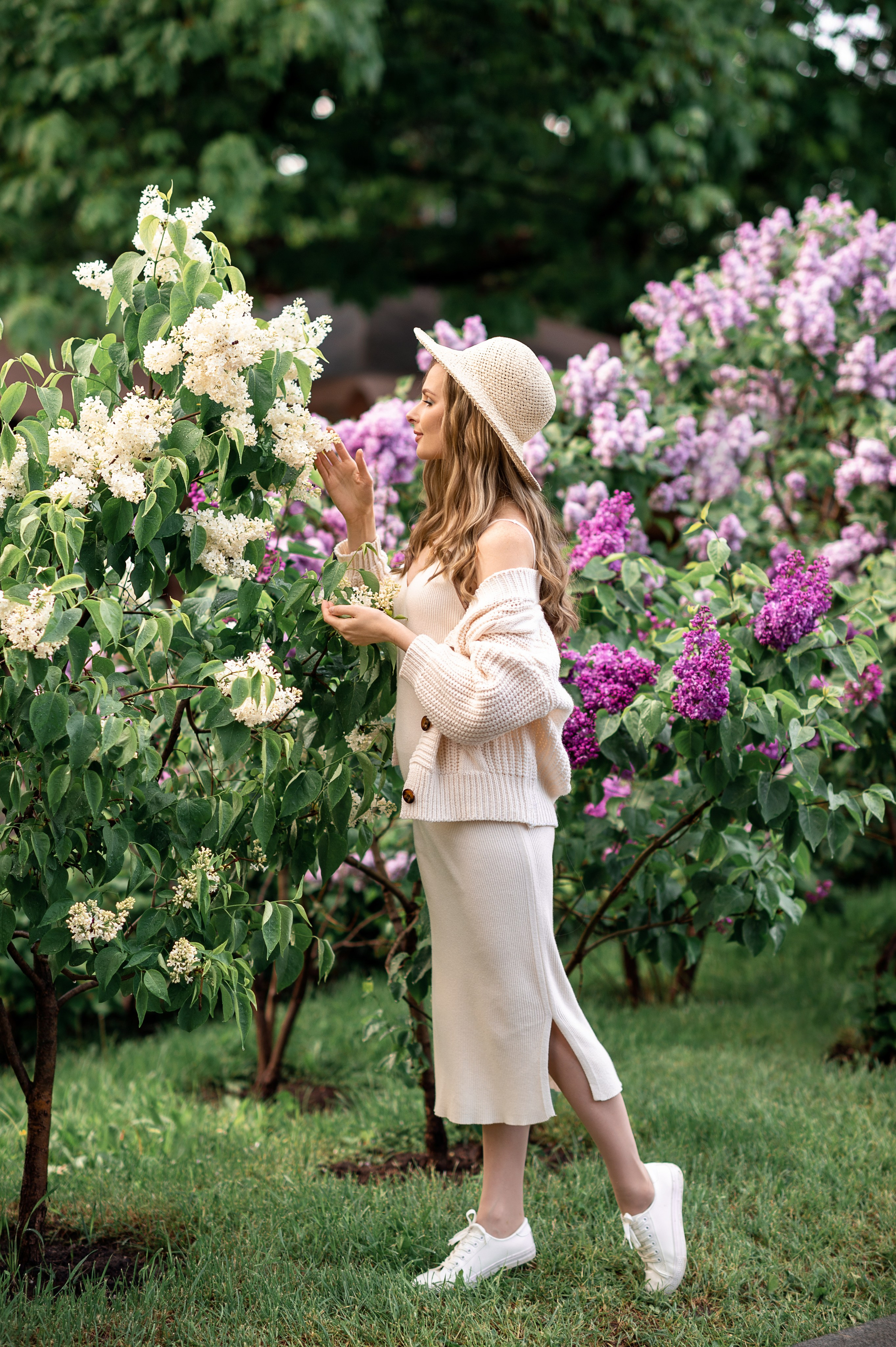 Молодая красивая девушка в светлой одежде и соломенной шляпке нюхает сирень