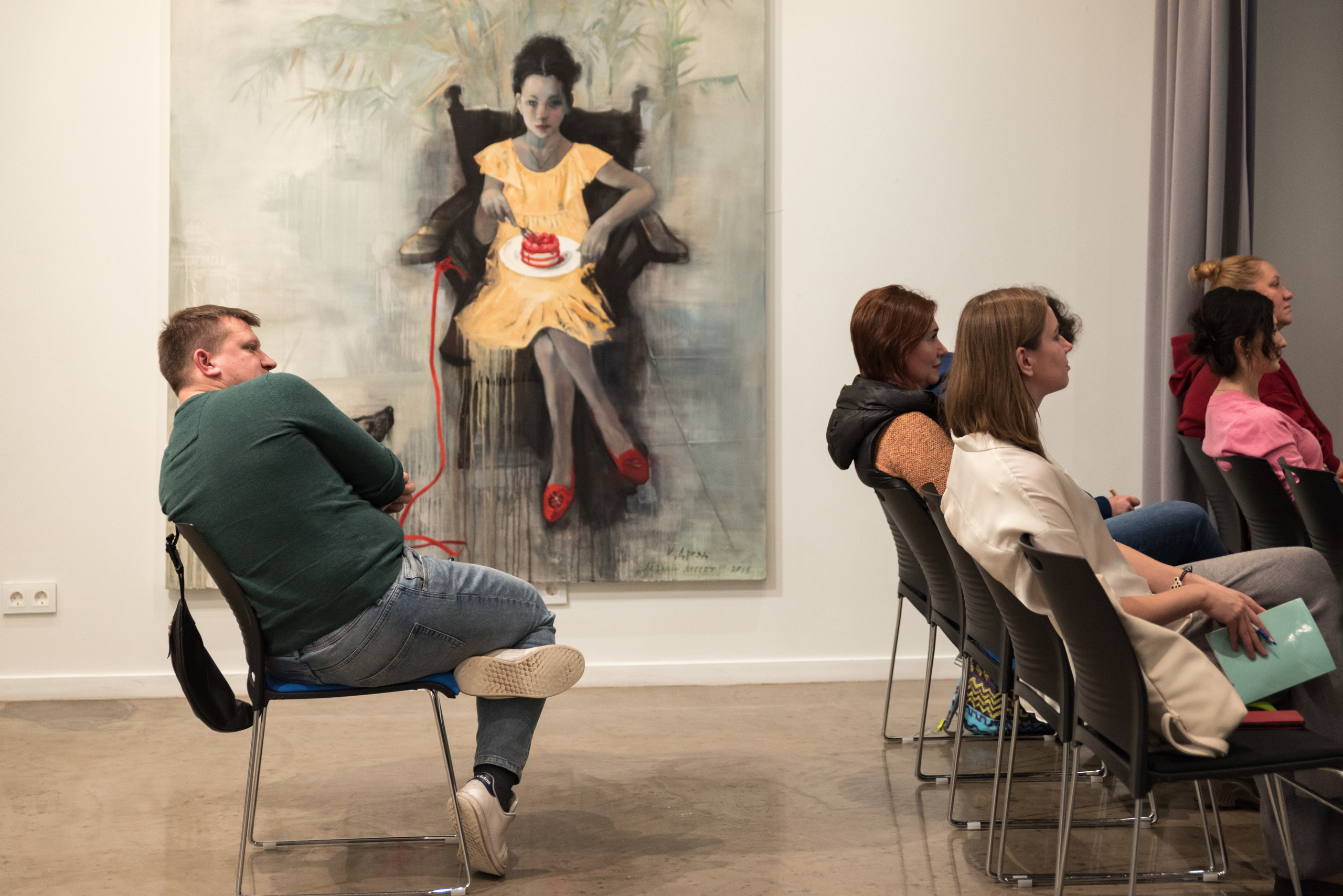 Мужчина сидит на стуле поджав одну ногу и откинувшись на спинку, за ним большая картина на стене.