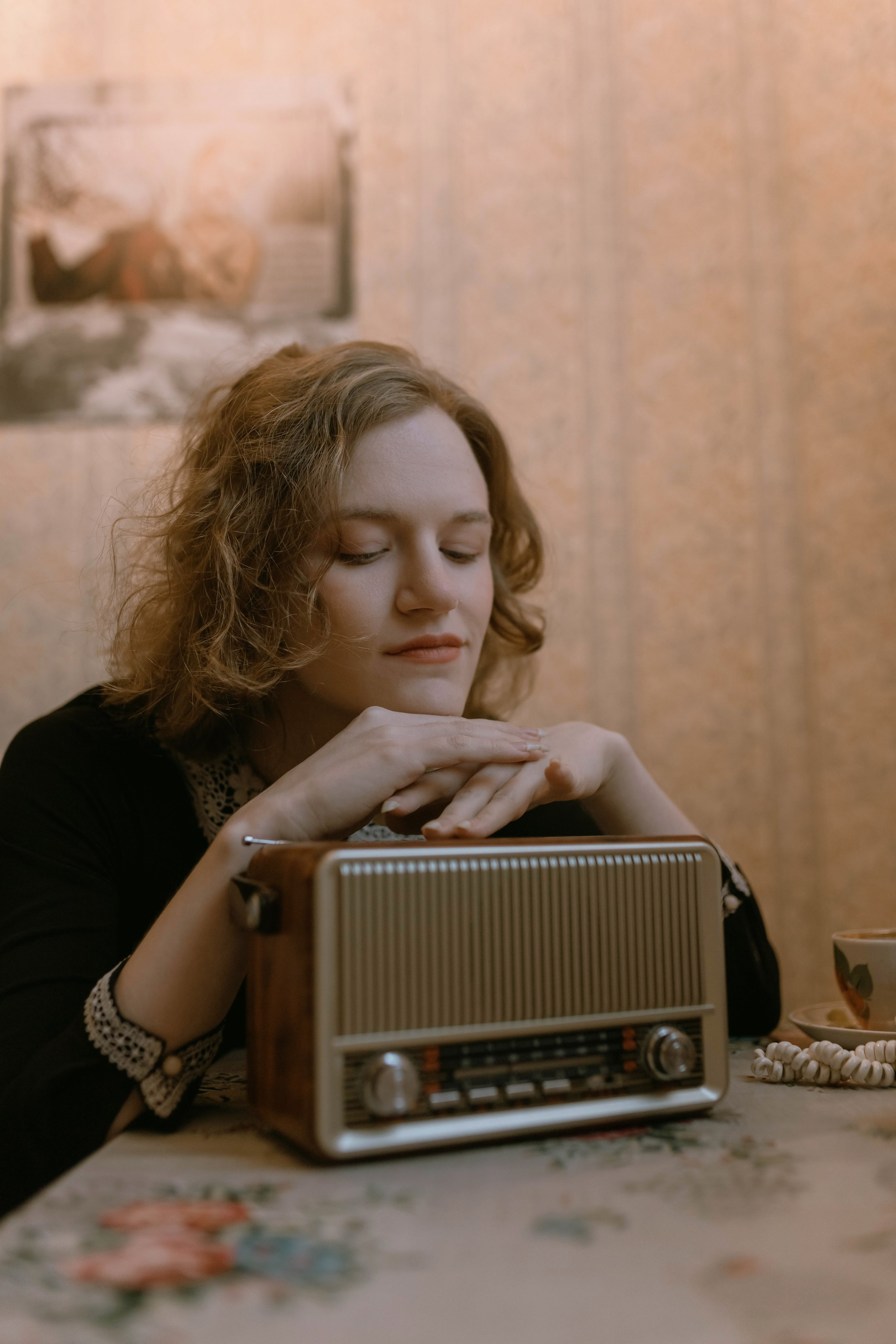 Молодая женщина с вьющимися волосами нежно подпирает подбородок руками, погрузившись в музыку, звучащую из старого радиоприемника. Теплая атмосфера комнаты усиливает ощущение расслабления.