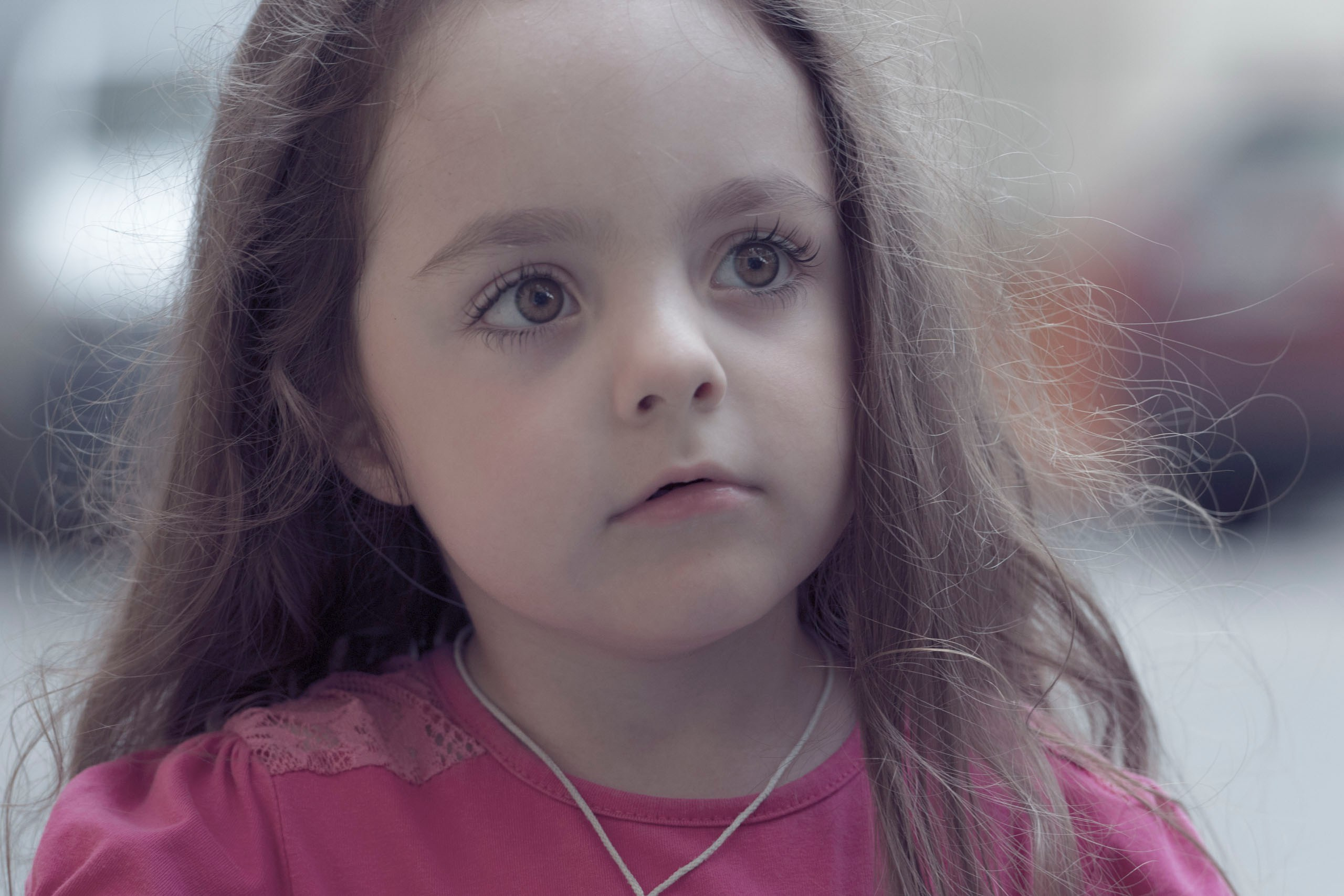 Крупный план портрета маленькой девочки с длинными каштановыми волосами и большими глазами. Она одета в розовую рубашку и смотрит вверх с задумчивым выражением лица.
