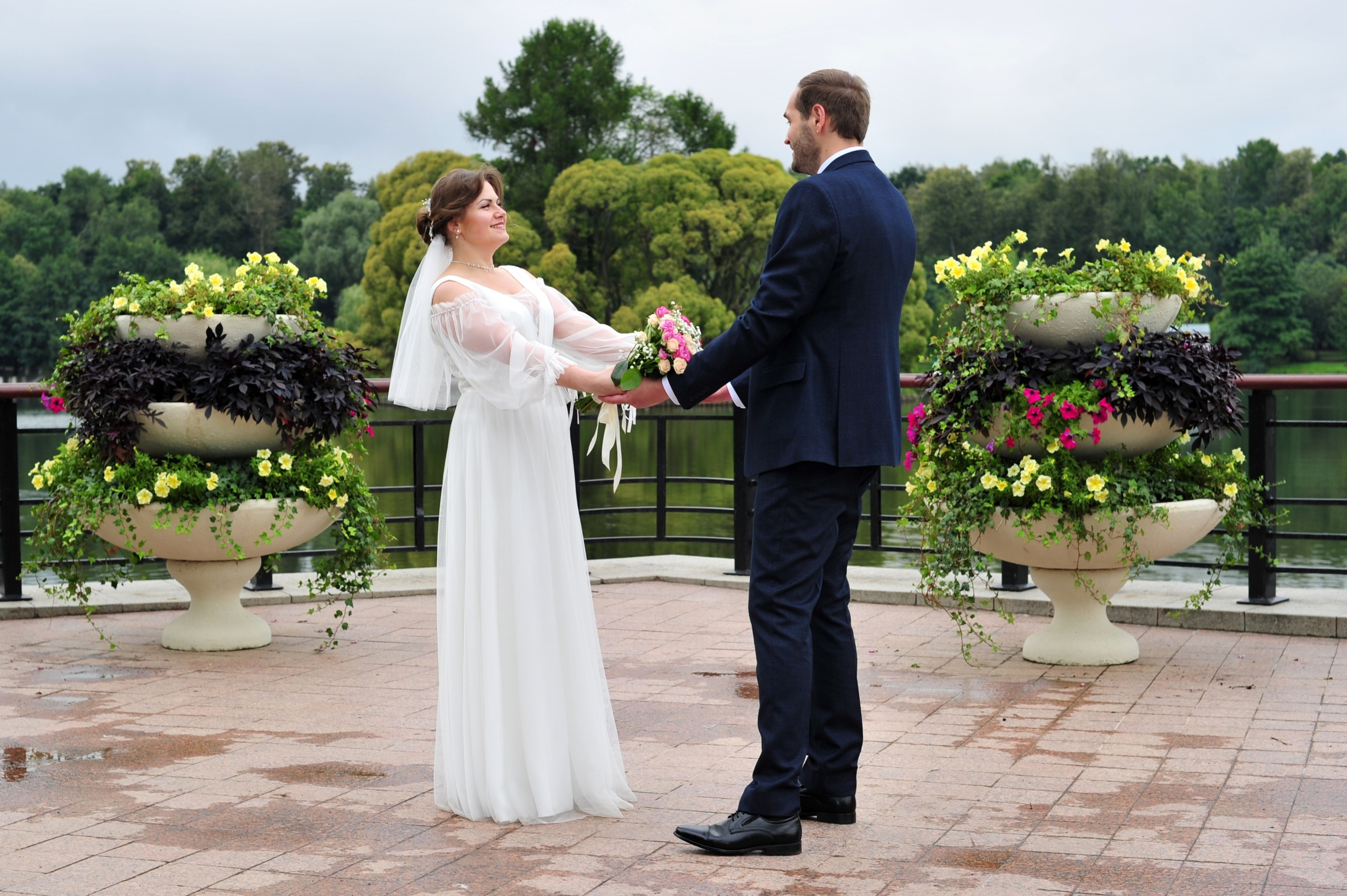 Свадьба в Царицыно пасмурным летним днем, новобрачные держатся за руки на фоне пруда и цветов