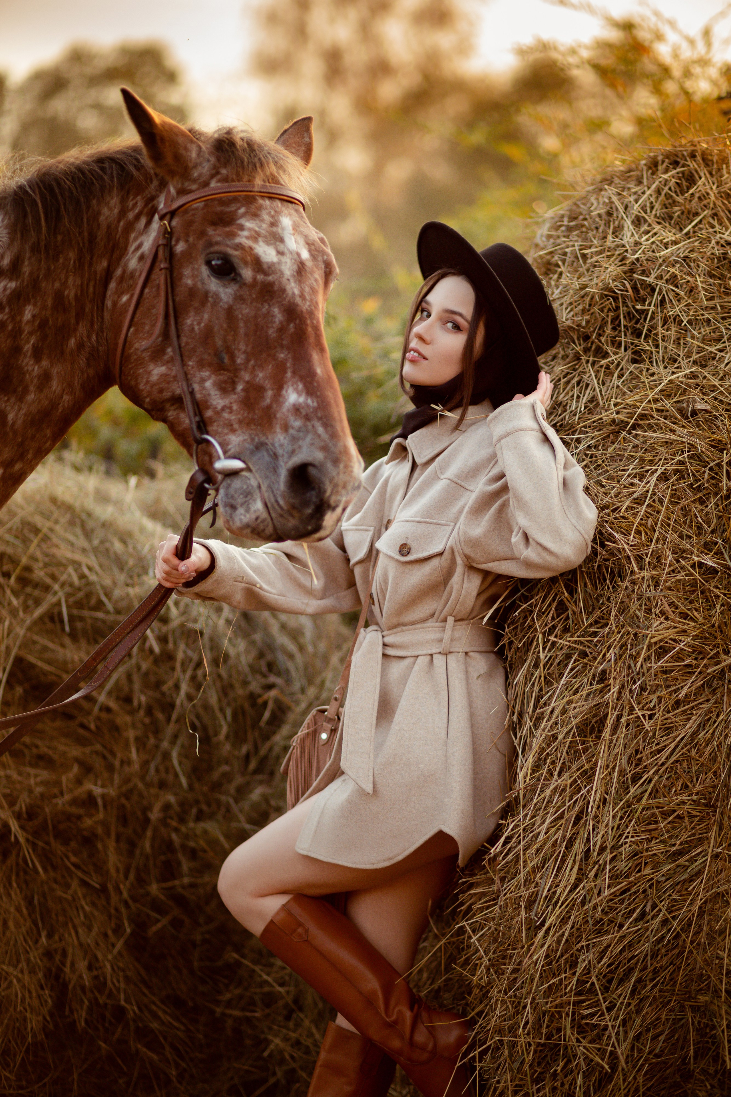 Девушка в черной шляпе стоит у сога сена и держит лошадь за поводья.