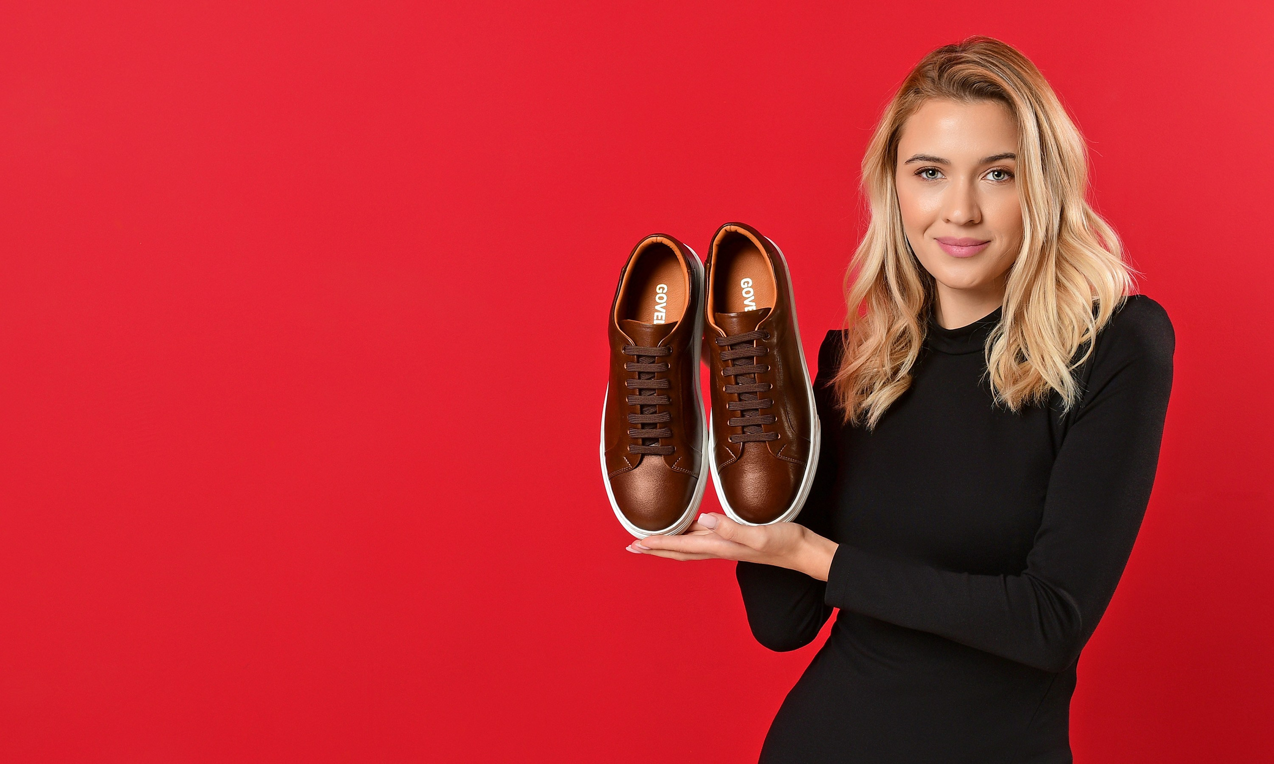 модель на красном фоне держит в руках кроссовки коричневого цвета