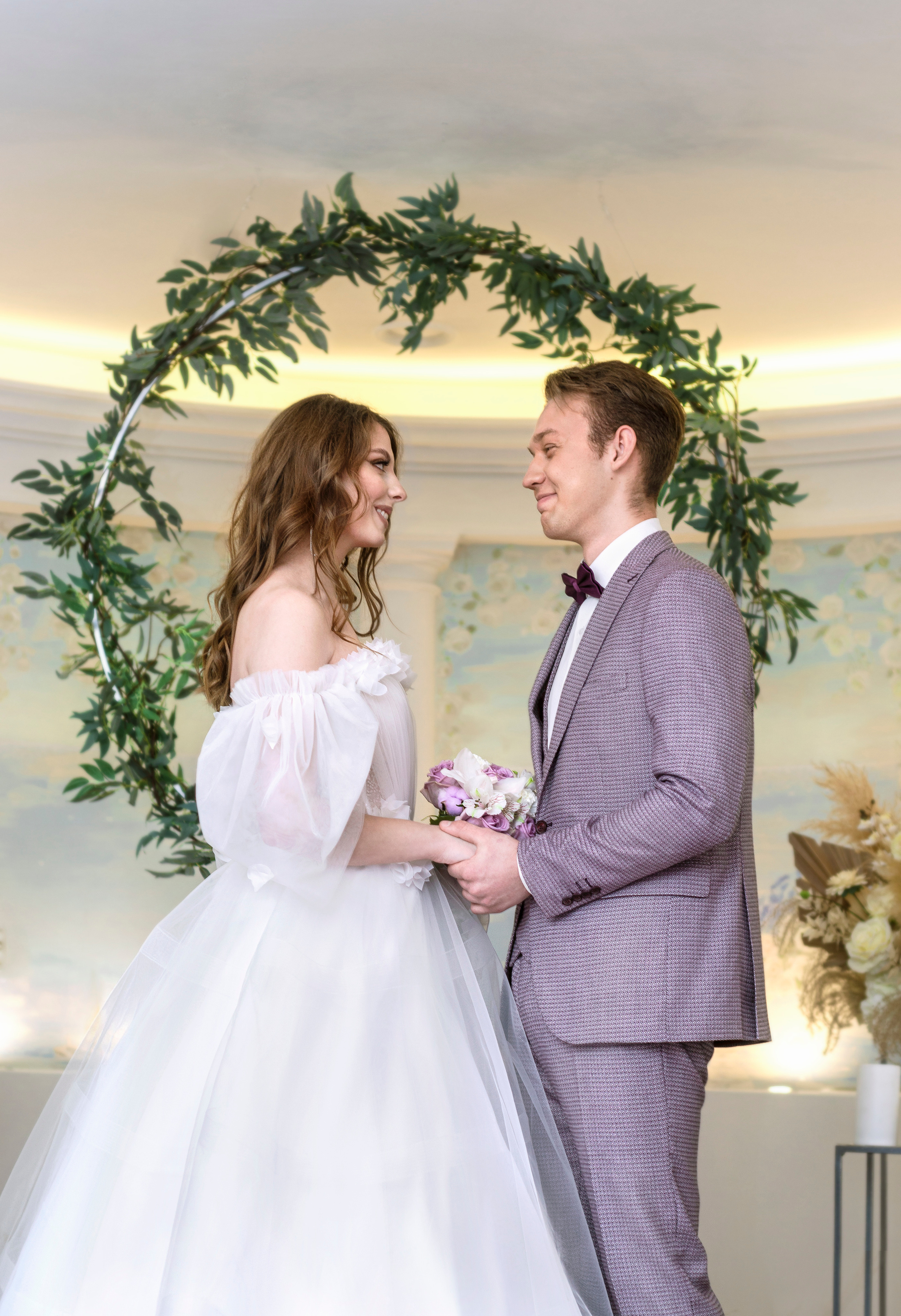 Взгляд жениха и невесты друг на друга во время свадебной церемонии на фоне зелёного кольца.