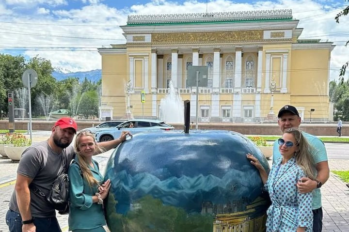 Вид на Лунный Каньон реки Чарын недалеко от Алматы, окружённый зелёными деревьями и кустарниками, на фоне красочных скал в солнечный день. На переднем плане женщина сидит на утёсе и любуется пейзажем