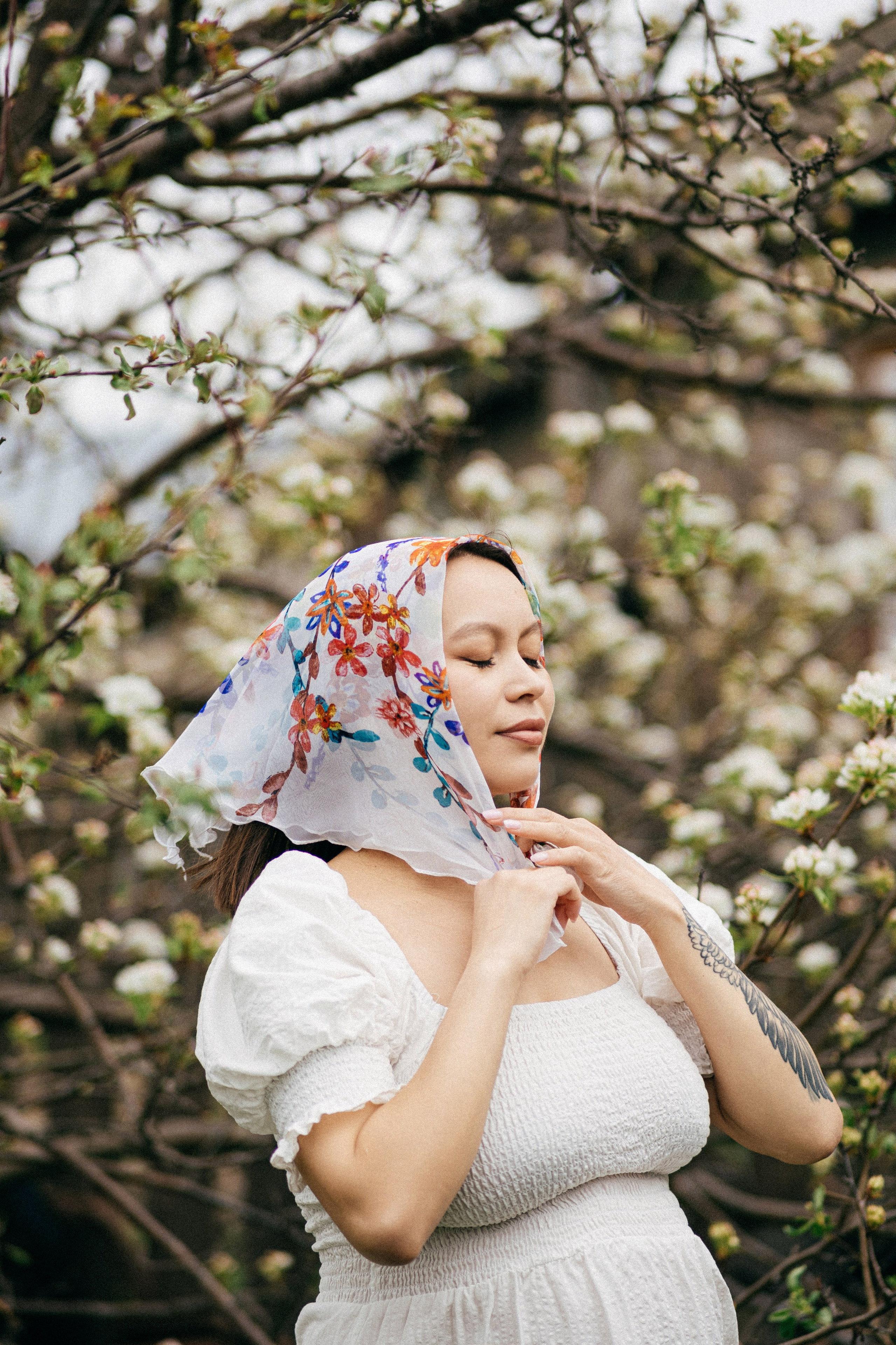 Беременная девушка в белом платье и косынке среди цветущих яблонь