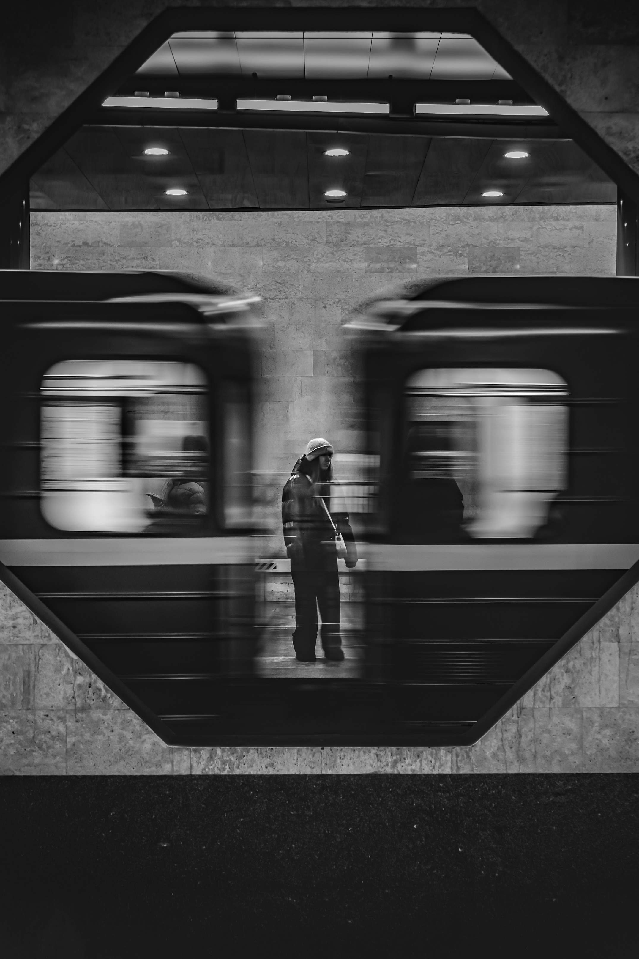 Одинокий человек в шапке стоит на платформе метро между двумя размытыми проезжающими поездами в октогональной раме. Стрит-фотография Мальцева Евгения.