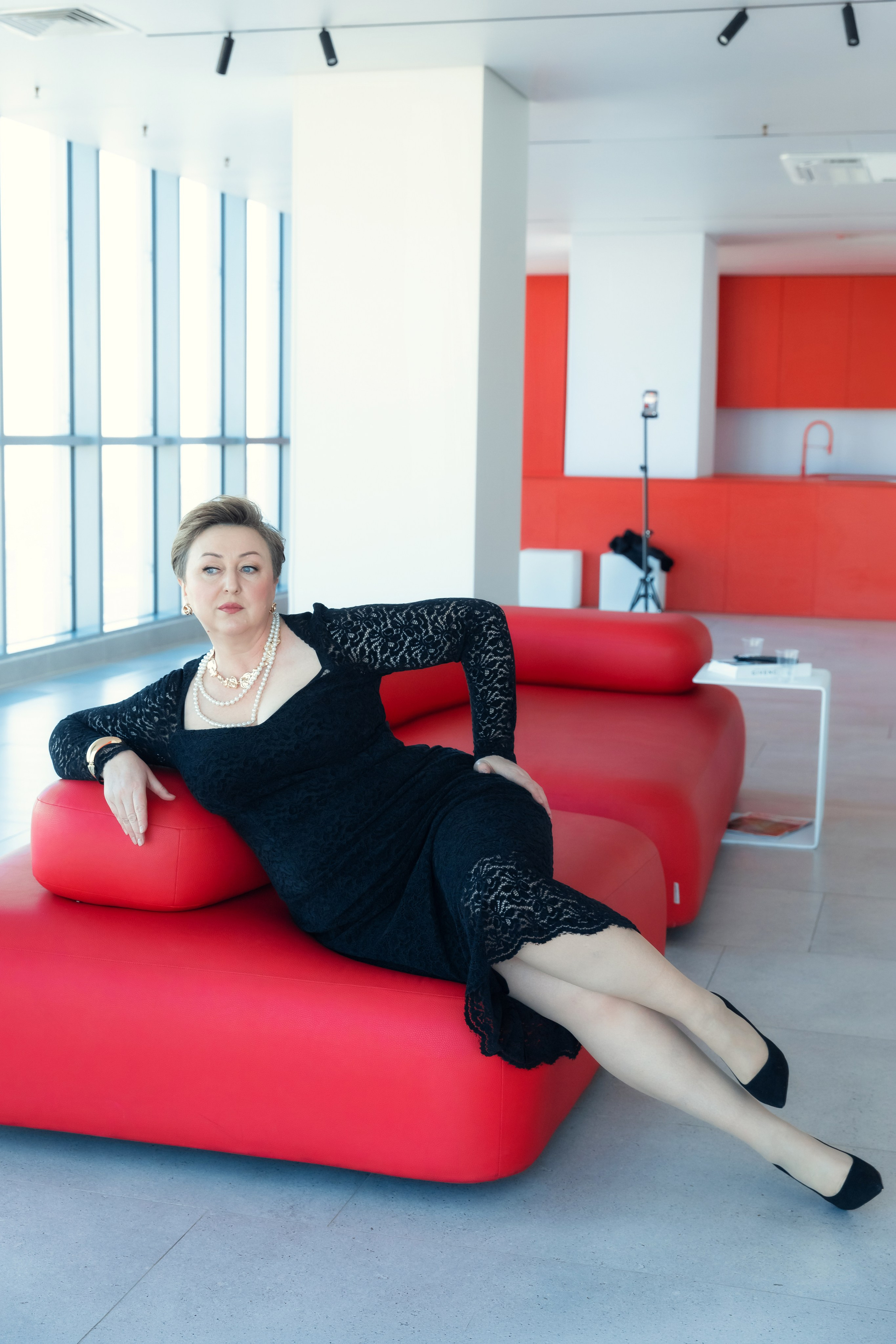 Женщина в черном кружевном платье лежит на красном диване в современном офисном интерьере, расслабленная поза