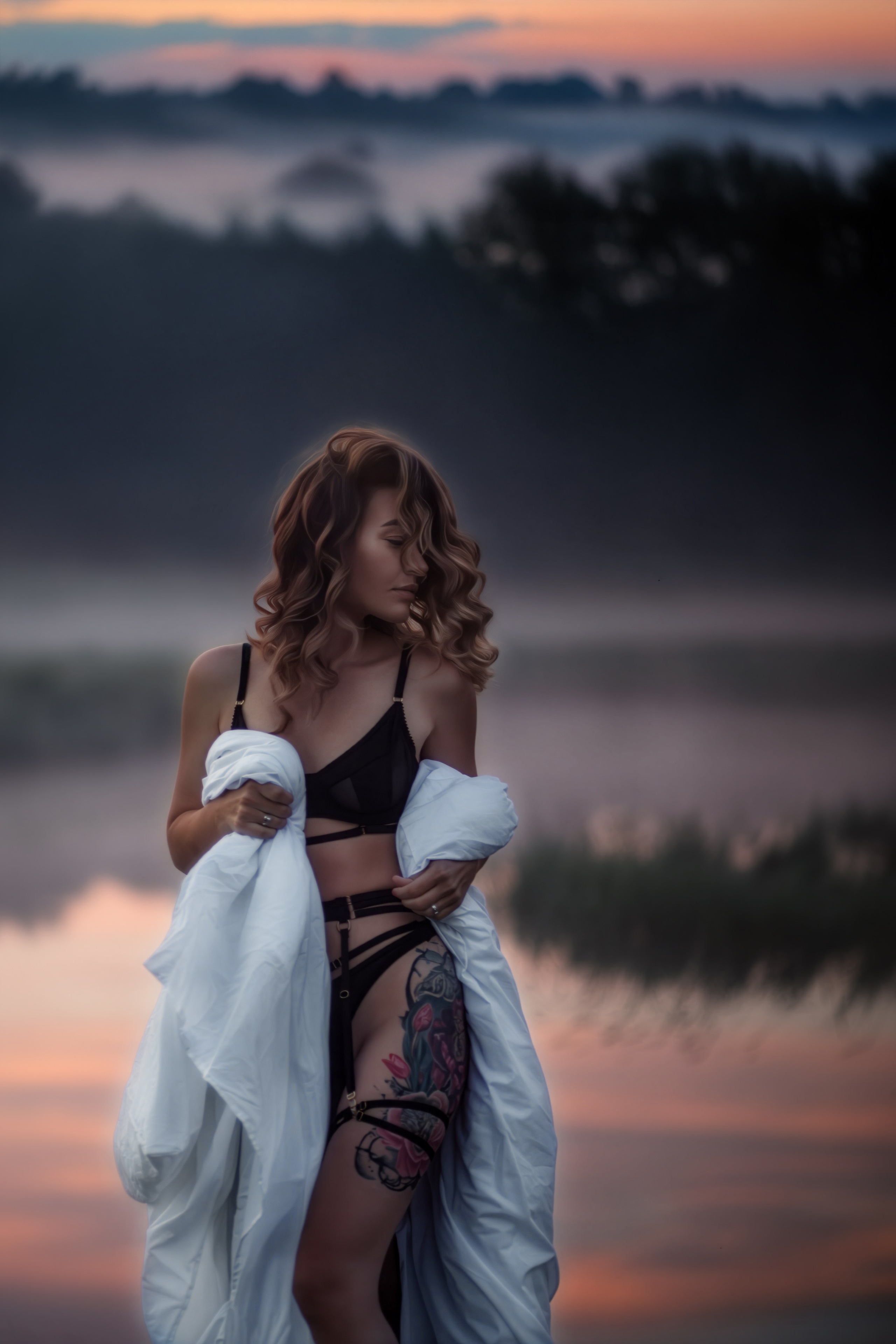 Девушка в чёрном белье и с татуировкой на бедре стоит у озера на рассвете, на плечах белая ткань, вокруг мягкий туман и отражение неба в воде.