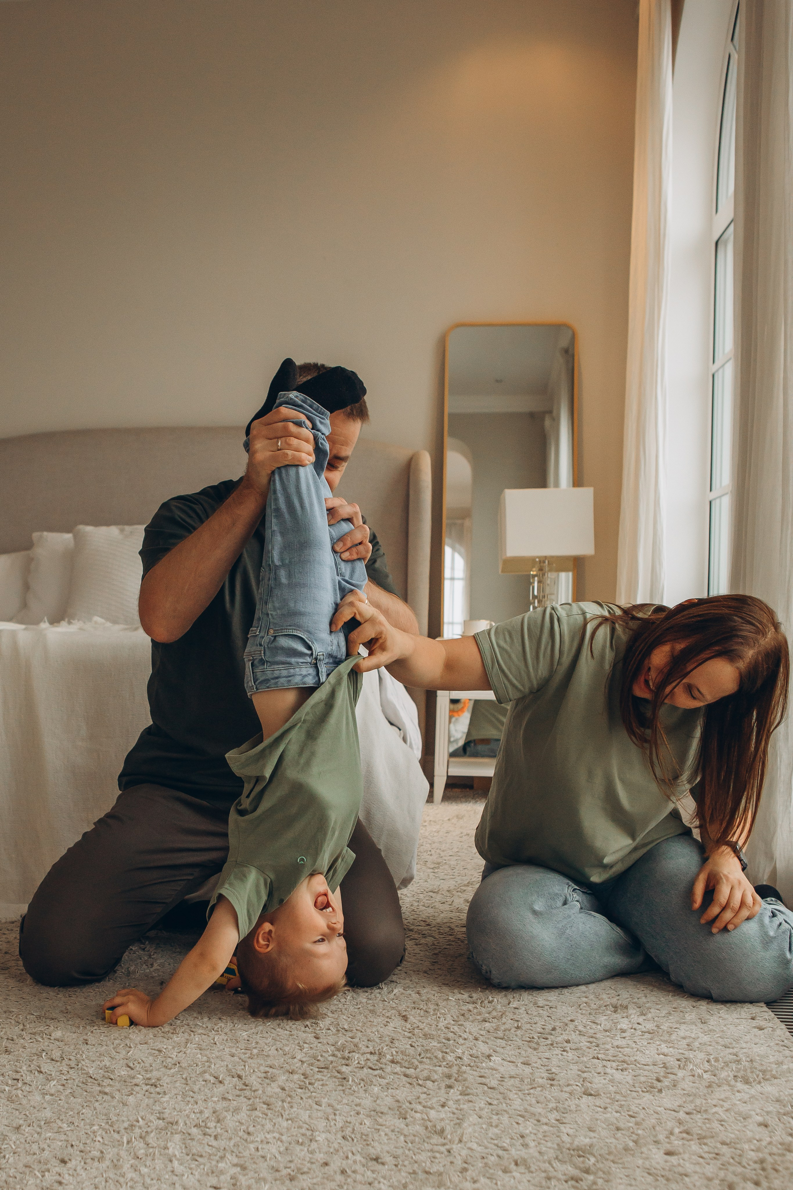 Семья игриво дурачится на ковре в светлом интерьере, папа держит счастливого ребенка за ноги. Детский и семейный фотограф Маруся Самылкина в Санкт-Петербурге.