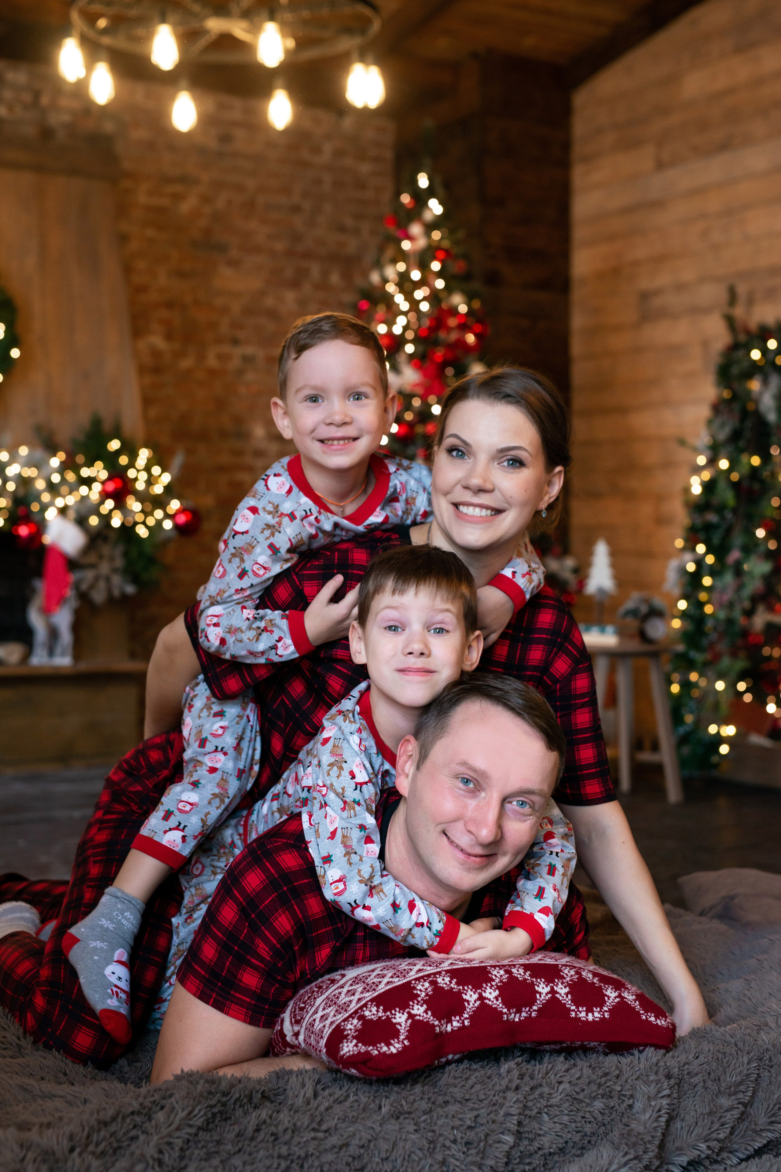 Молодые и озорные мама и папа со своими двумя сыновьями в красных пижамах, обнимая друг друга и взвалившись на плечи главы семейства, с радостью позируют во время студийной семейной новогодней фотосессии в Санкт-Петербурге.