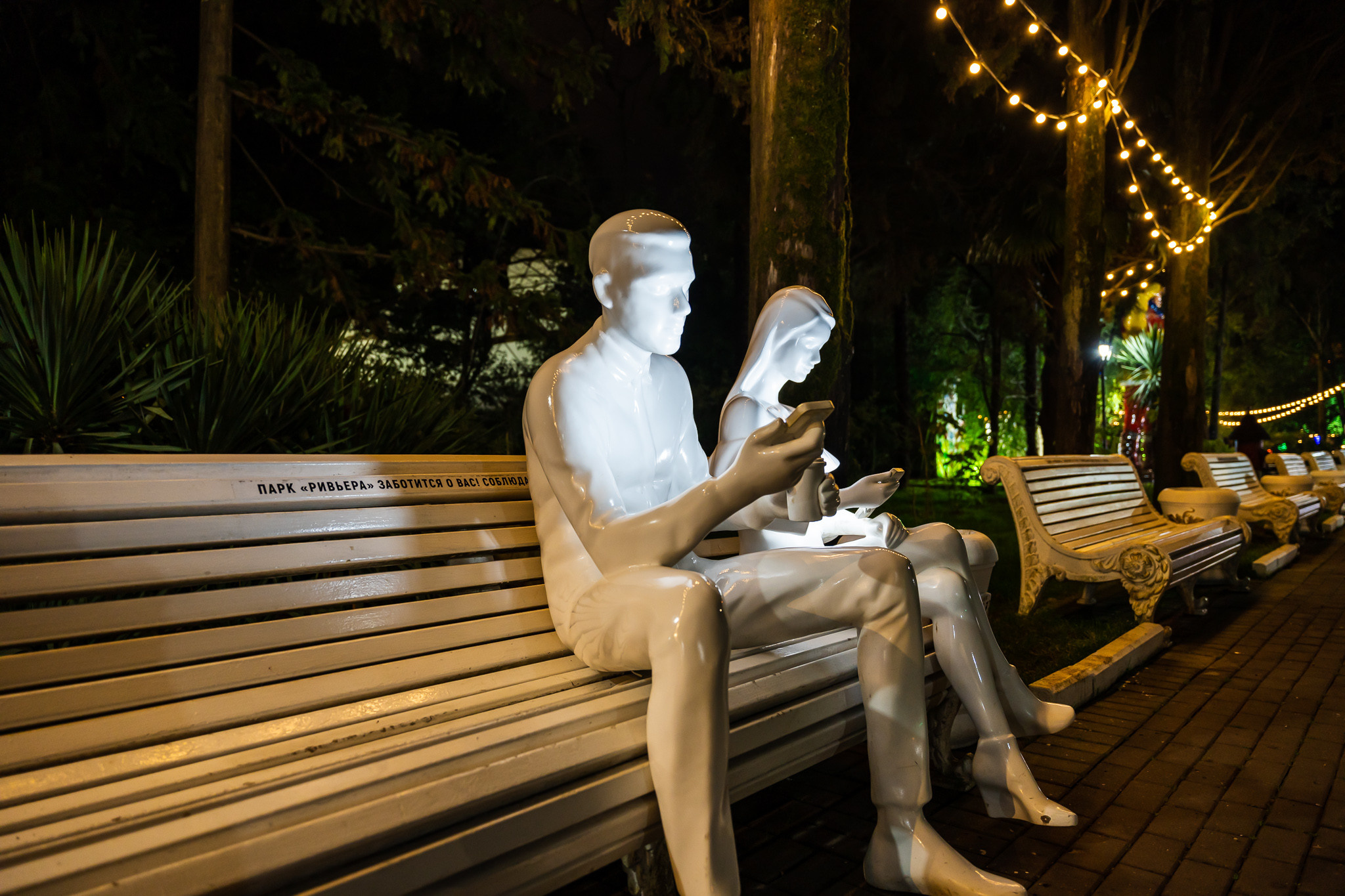 Фигуры людей с мобильными телефонами, сидящих на скамейке в парке Ривьера в Сочи ночью с подсветкой 