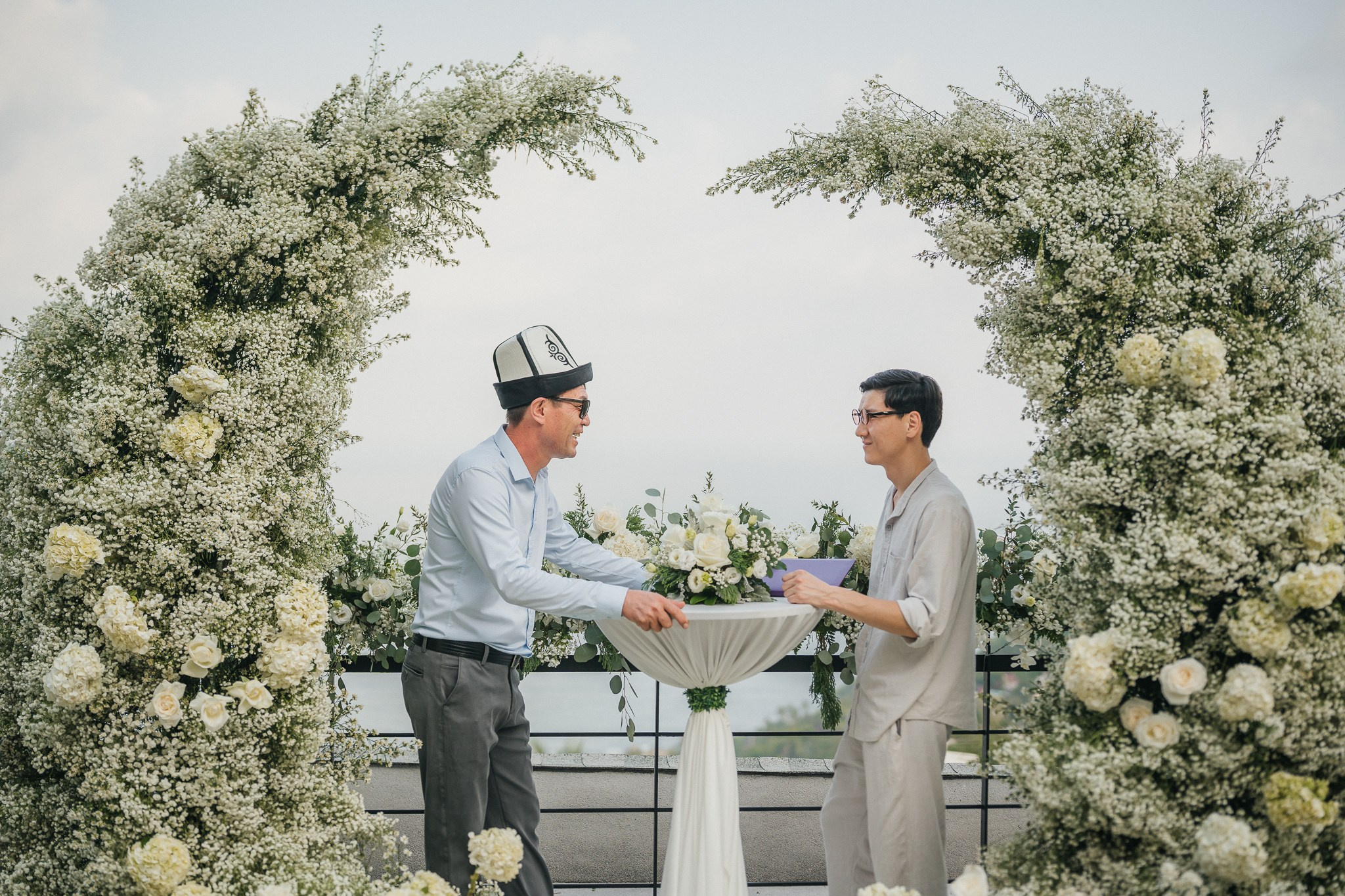 Two men exchanging vows during an intimate beach wedding ceremony beneath a lush white floral arch overlooking the ocean in Koh Samui, Thailand.