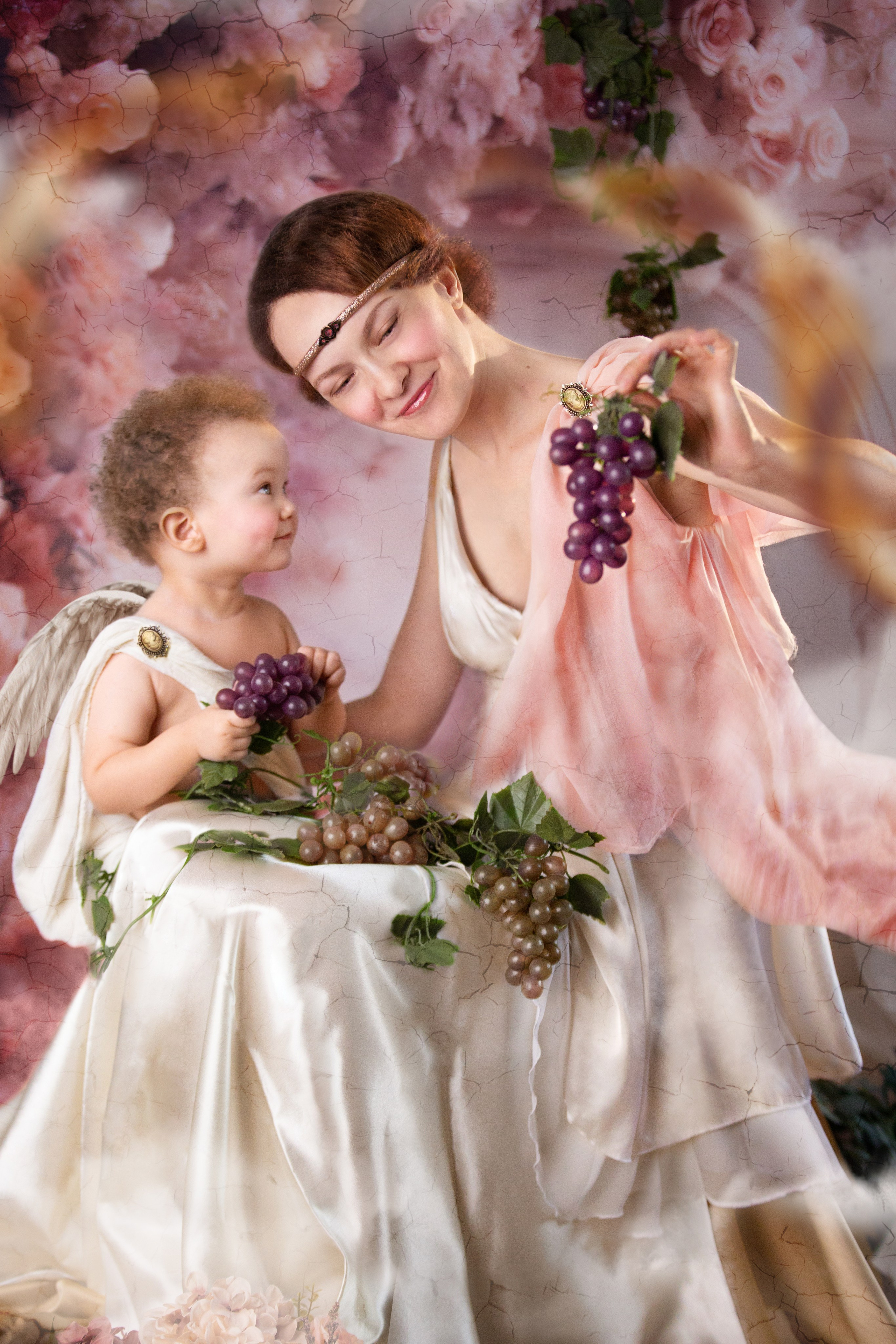 Женщина и ребенок в ангельских одеяниях в древнегреческом стиле держат в руках виноградные гроздья, окружены ботаническим фоном с розовыми цветами и зелеными листьями. Малыш восторженно смотрит на маму.