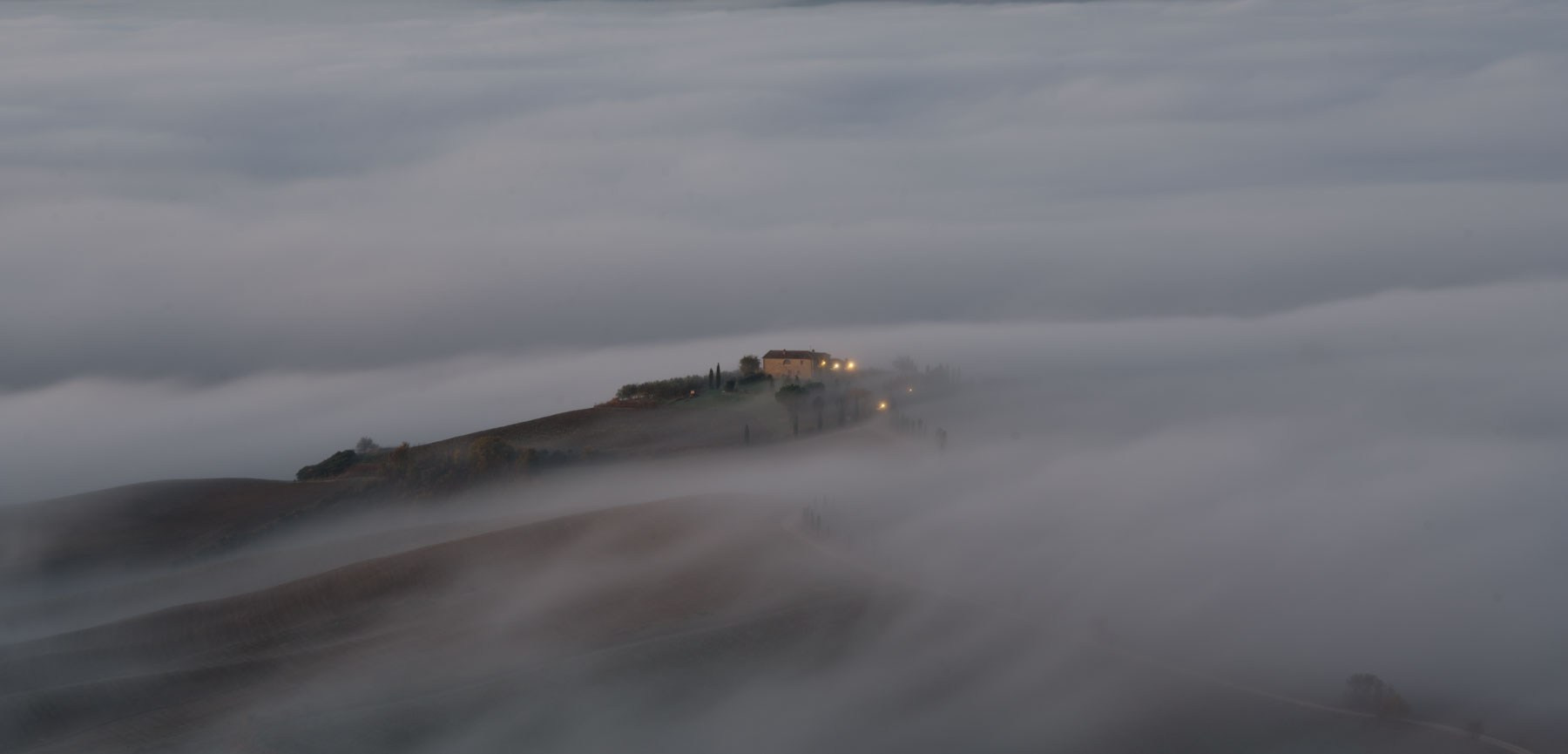 «Утро в сердце Тосканы»Завораживающий снимок туманной Тосканы на рассвете, передающий атмосферу покоя и умиротворения. На переднем плане — волнистые, вспаханные поля в теплых оттенках утреннего света. Извилистая дорога, обрамленная стройными кипарисами, ведет к уединенной тосканской вилле (Podere Belvedere) на вершине холма. Мягкая дымка тумана окутывает дальние холмы, создавая многослойный, художественный пейзаж, характерный для региона Валь-д'Орча (Val d'Orcia). Эта картина — воплощение классического итальянского сельского вида, известного во всем мире.