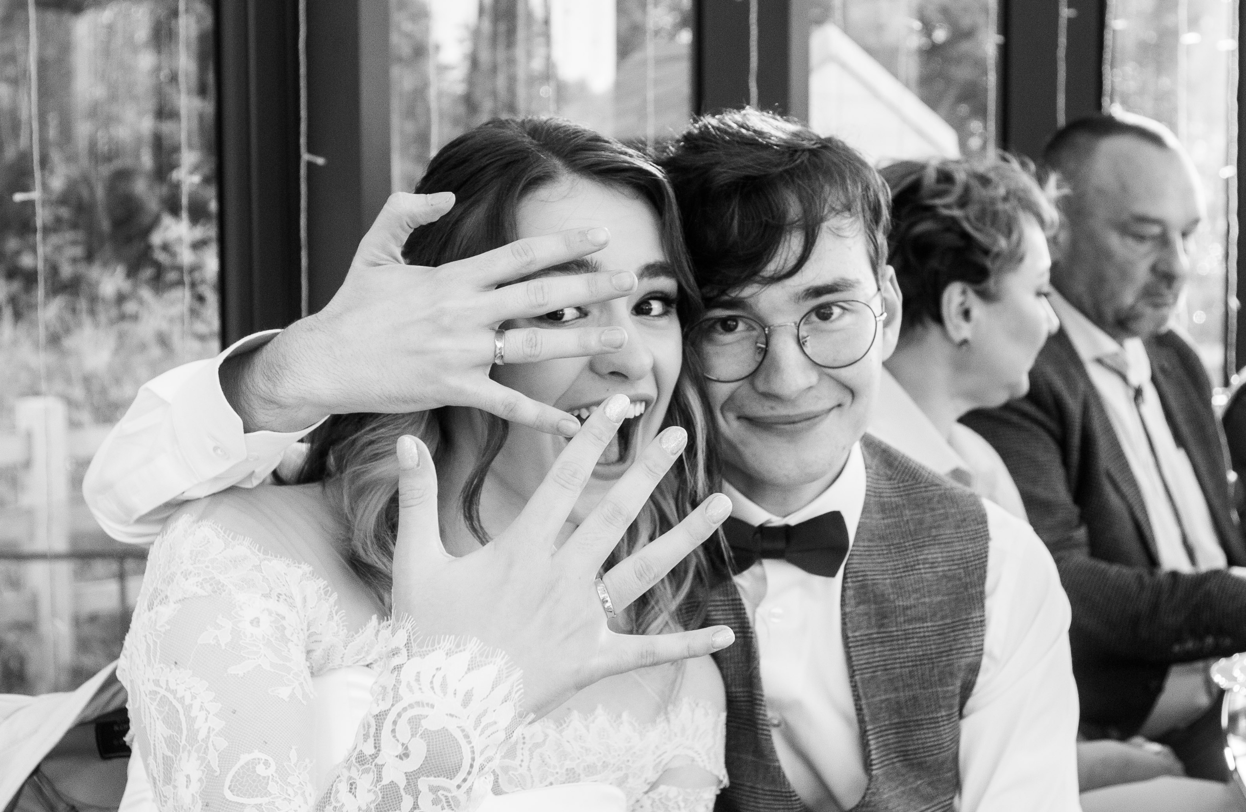 Жених и невеста веселятся и показывают пальцы с кольцами на свадебном банкете, фото ч/б, фотограф видеооператор Павел Коротков