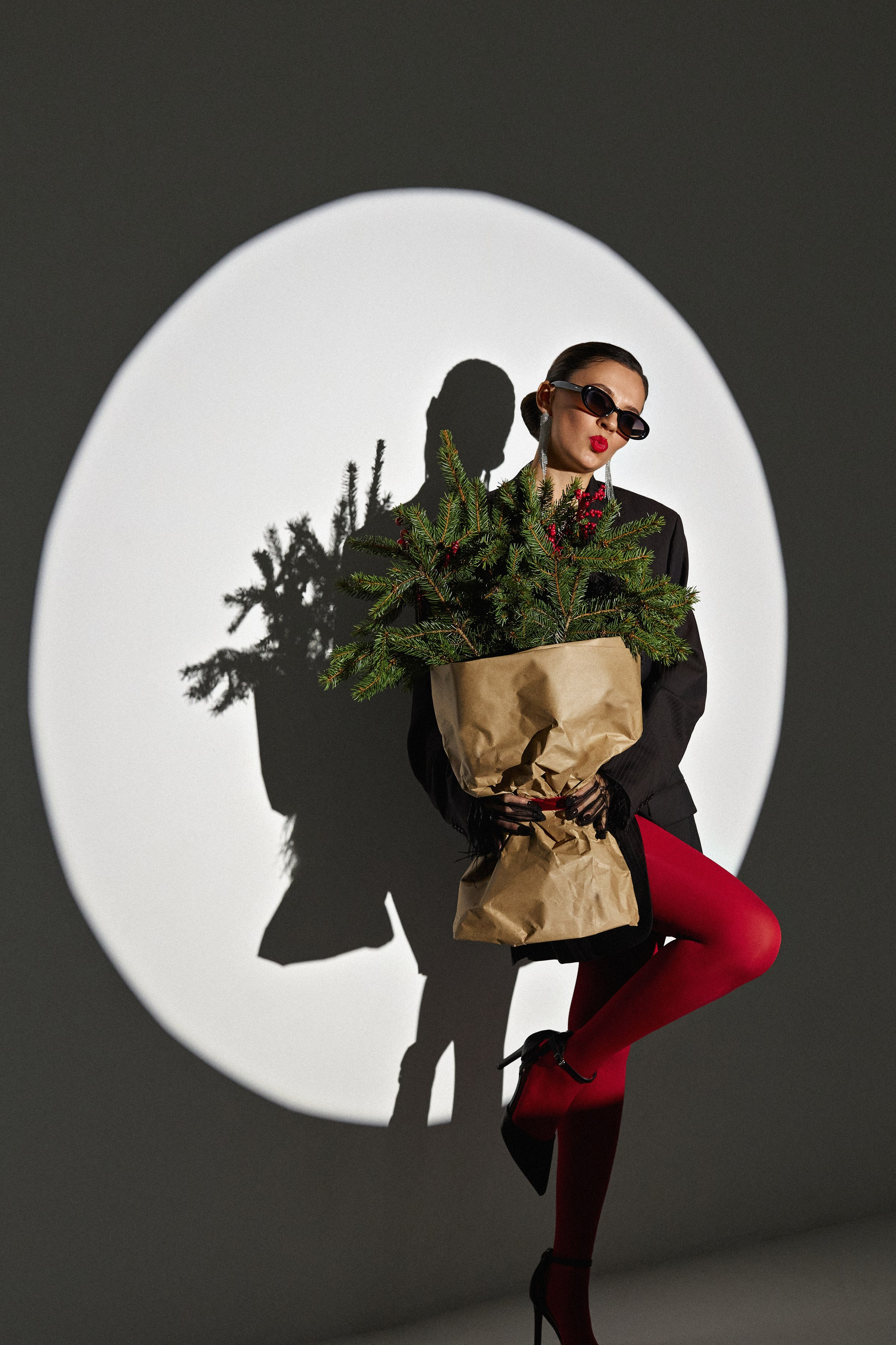 Красивая девушка с пучком на голове в черном пиджаке c перьями на рукавах и красных колготках на новогодней фотосессии в студии с букетом из еловых веток в колье в минималистичном стиле в световом круге в Домодедово в фотостудии LUMI
