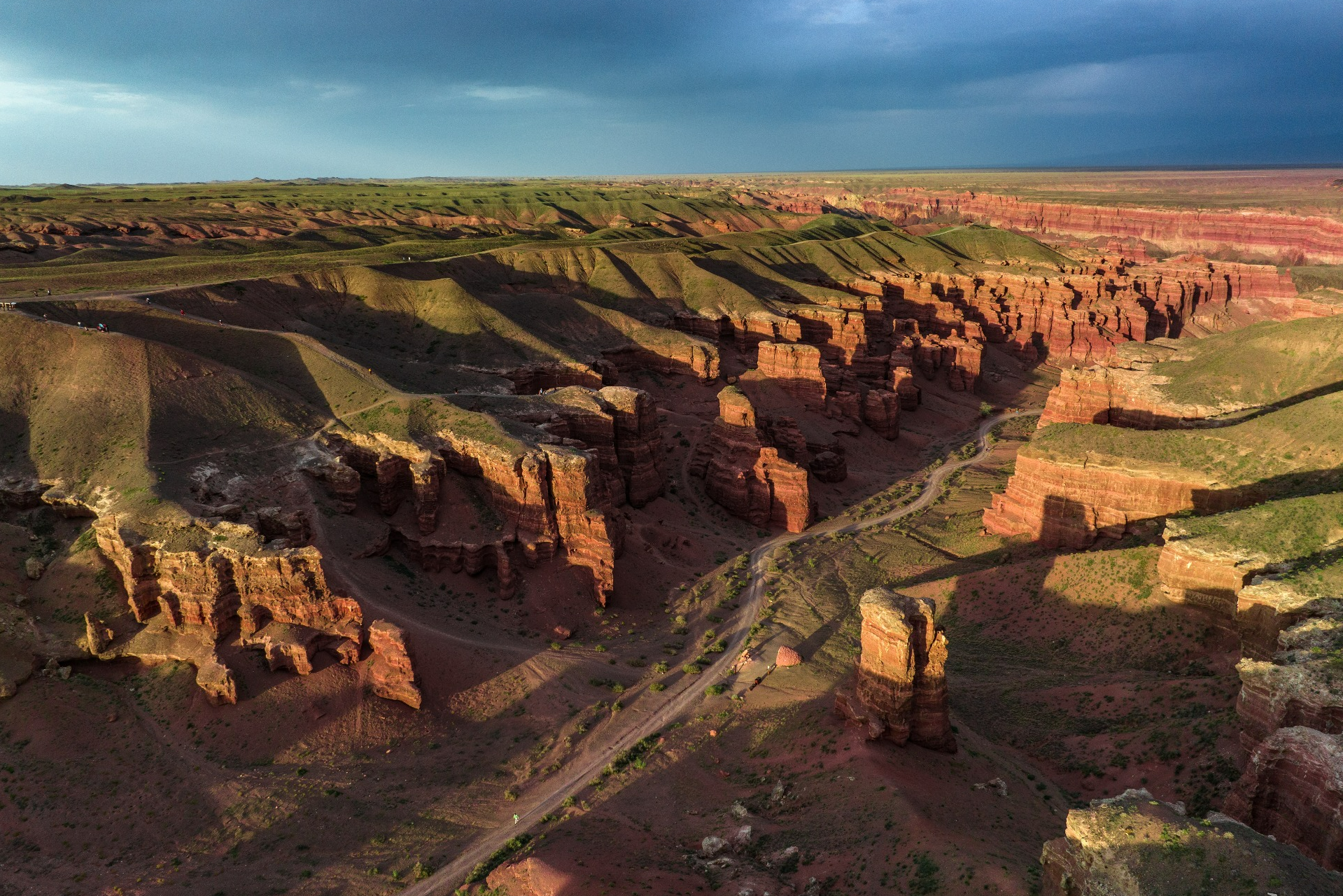Величественный Чарынский каньон в районе Алматы с ярко-красными скалами, пересечённый извилистой дорогой, на которую падают длинные вечерние тени. Захватывающее сочетание красочных утёсов, зелёных склонов и драматичного неба создаёт эффектный пейзаж