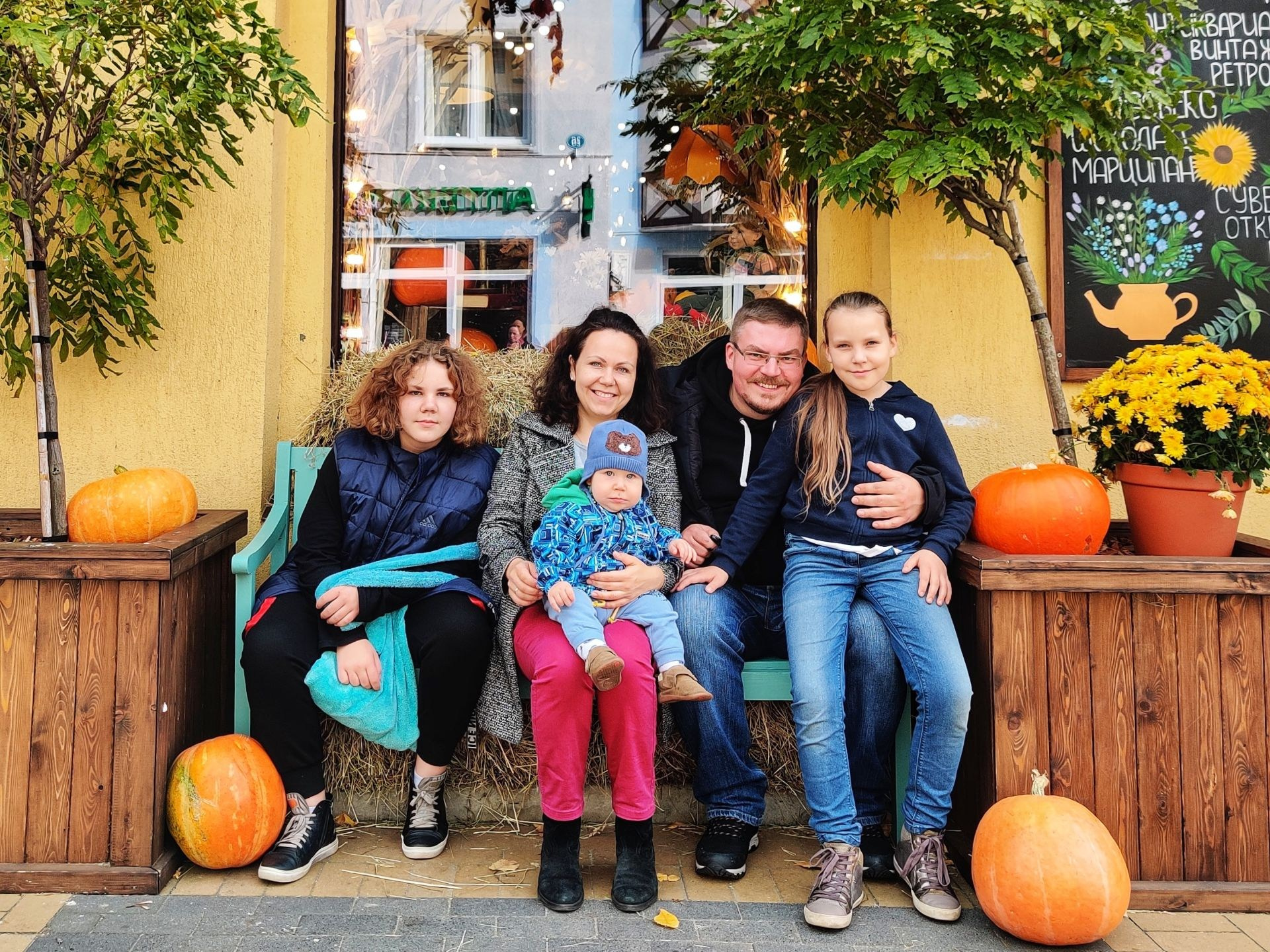 Семья с детьми сидит на скамейке с тыквами у входа в кафе