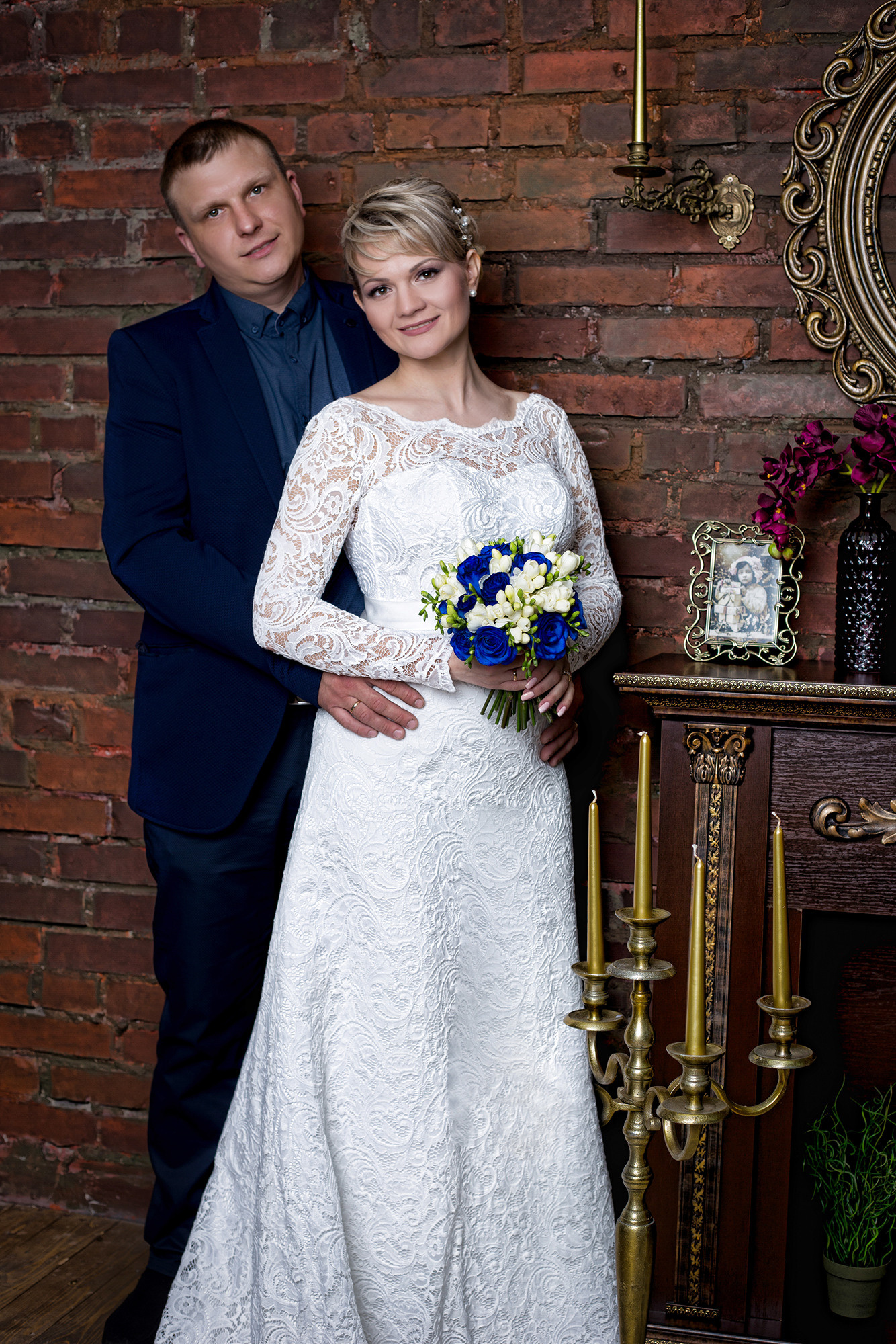 жених в темно-синем костюме и невеста в белом длинном закрытом платье на фото камина и кирпичной стены