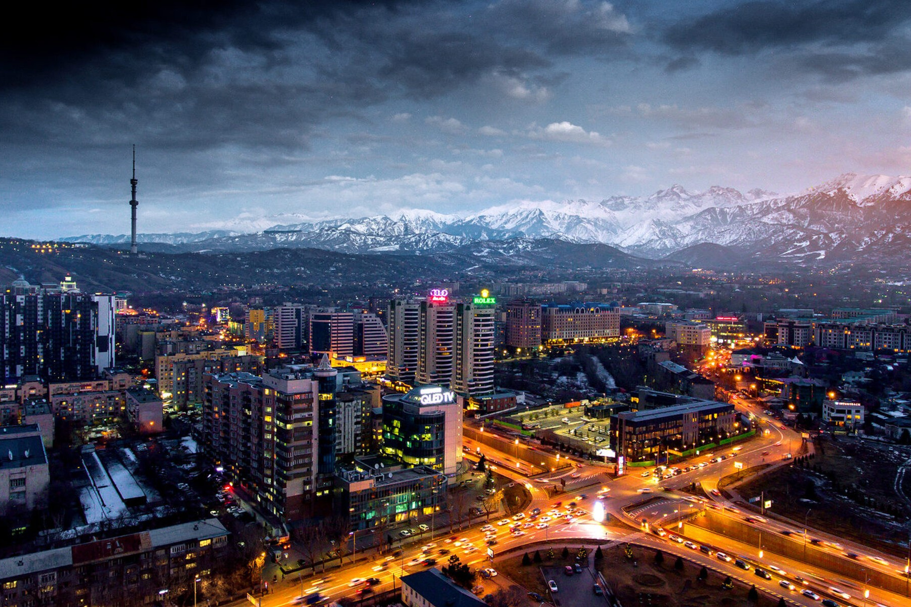 Панорамный вид на ночной город Алматы в Казахстане с высоты. В центре кадра сияют огни зданий, улицы заполнены светом автомобилей. На горизонте виднеются величественные заснеженные горы, создавая контраст с урбанистическим пейзажем. В левой части кадра возвышается телевизионная башня, подчеркивающая городской ландшафт. Облака в небе добавляют драматичности сцене.