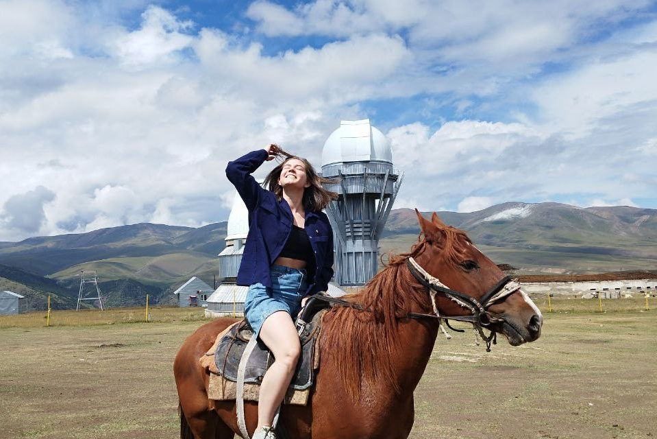 Величественный Чарынский каньон в районе Алматы с ярко-красными скалами, пересечённый извилистой дорогой, на которую падают длинные вечерние тени. Захватывающее сочетание красочных утёсов, зелёных склонов и драматичного неба создаёт эффектный пейзаж