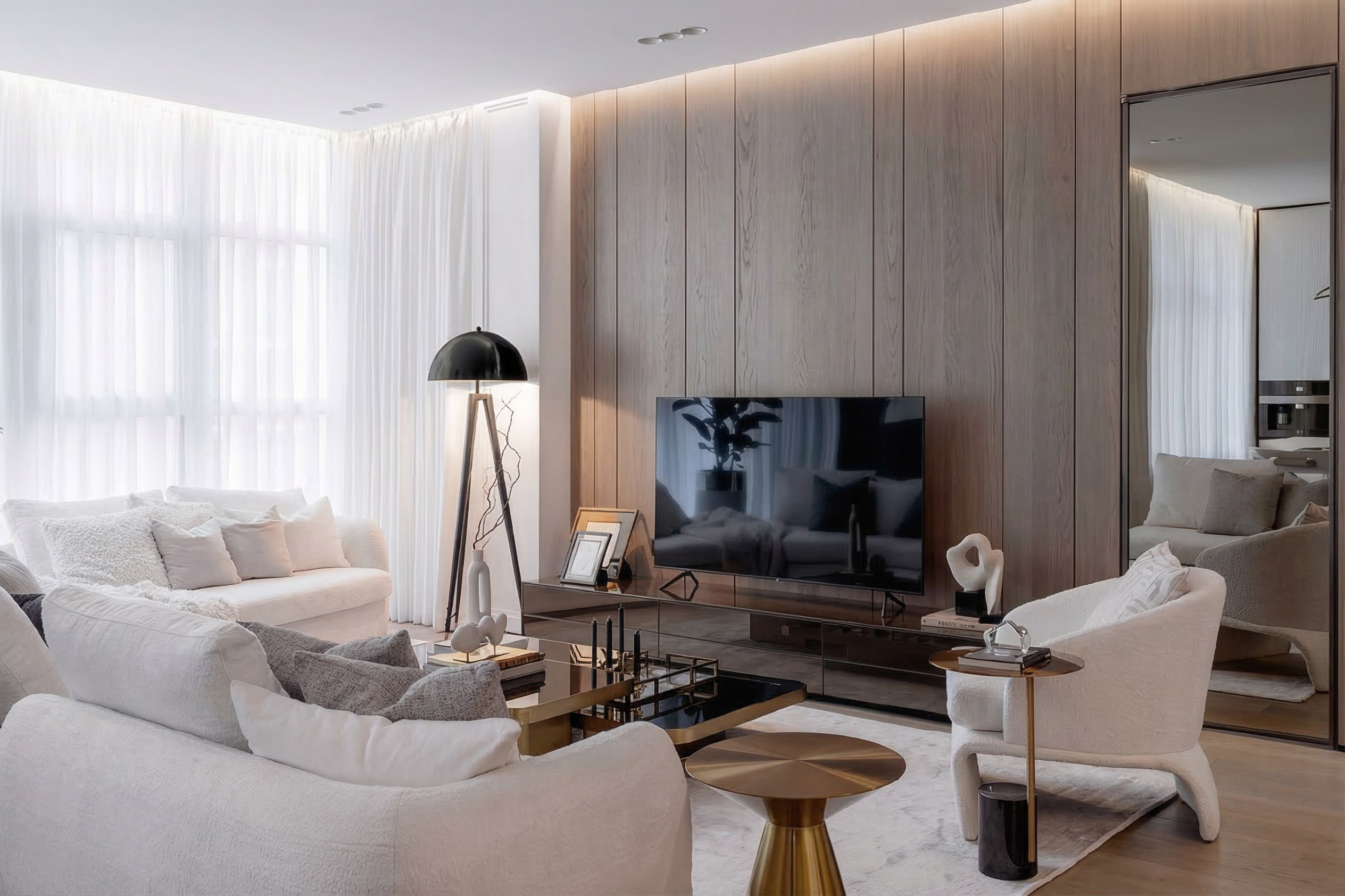 Светлый интерьер гостиной с деревянной отделкой стен, белым диваном, креслом букле и большим зеркалом. В центре — ТВ-зона и золотистые журнальные столики, у окна стоит черный дизайнерский торшер на треноге.