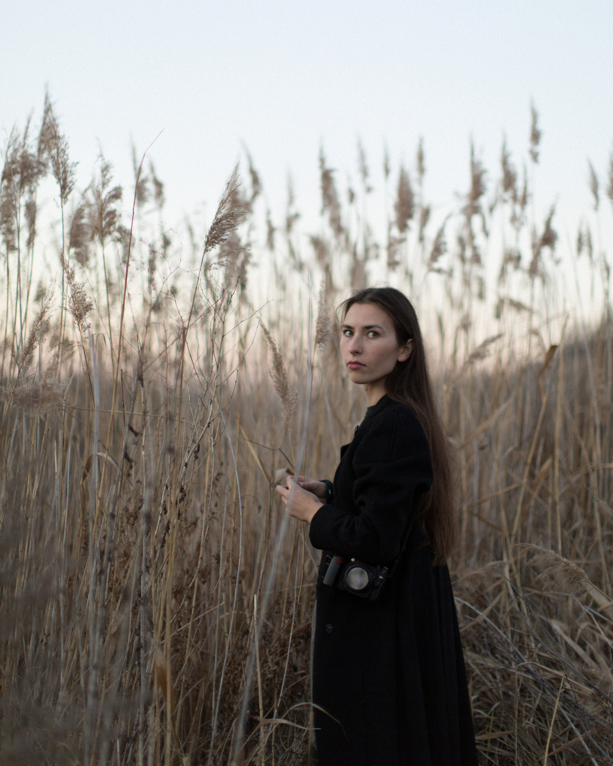 Уличный портрет девушки в черном пальто с фотоаппаратом среди камышей.